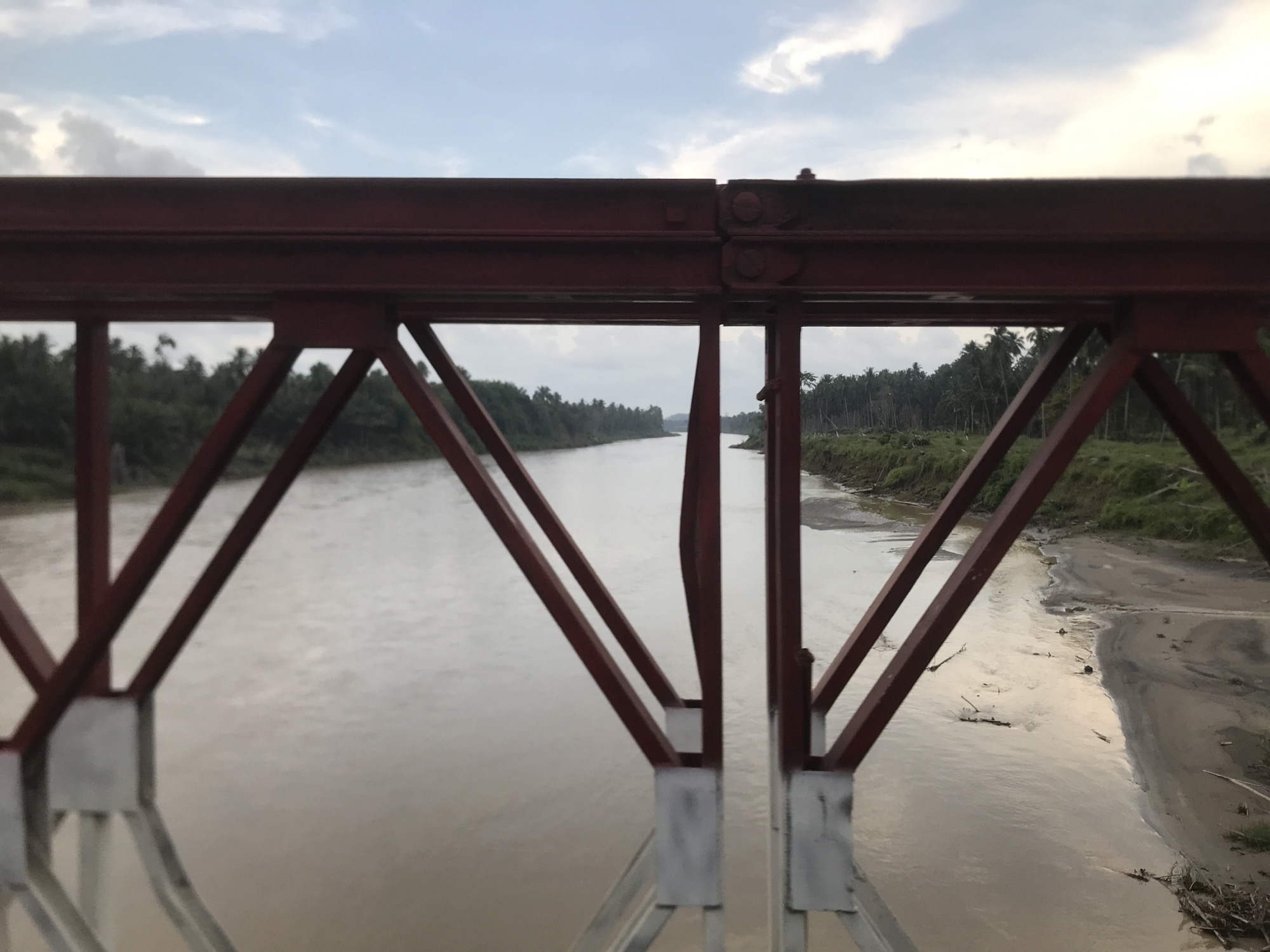 The image shows a view from a red metal bridge in Awegeutahpaya, Indonesia. The bridge's framework is visible in the foreground. Beyond the bridge structure is a wide, murky river flowing away into the distance. The riverbanks are lined with green vegetation, including trees and some grassy areas. The sky is overcast.