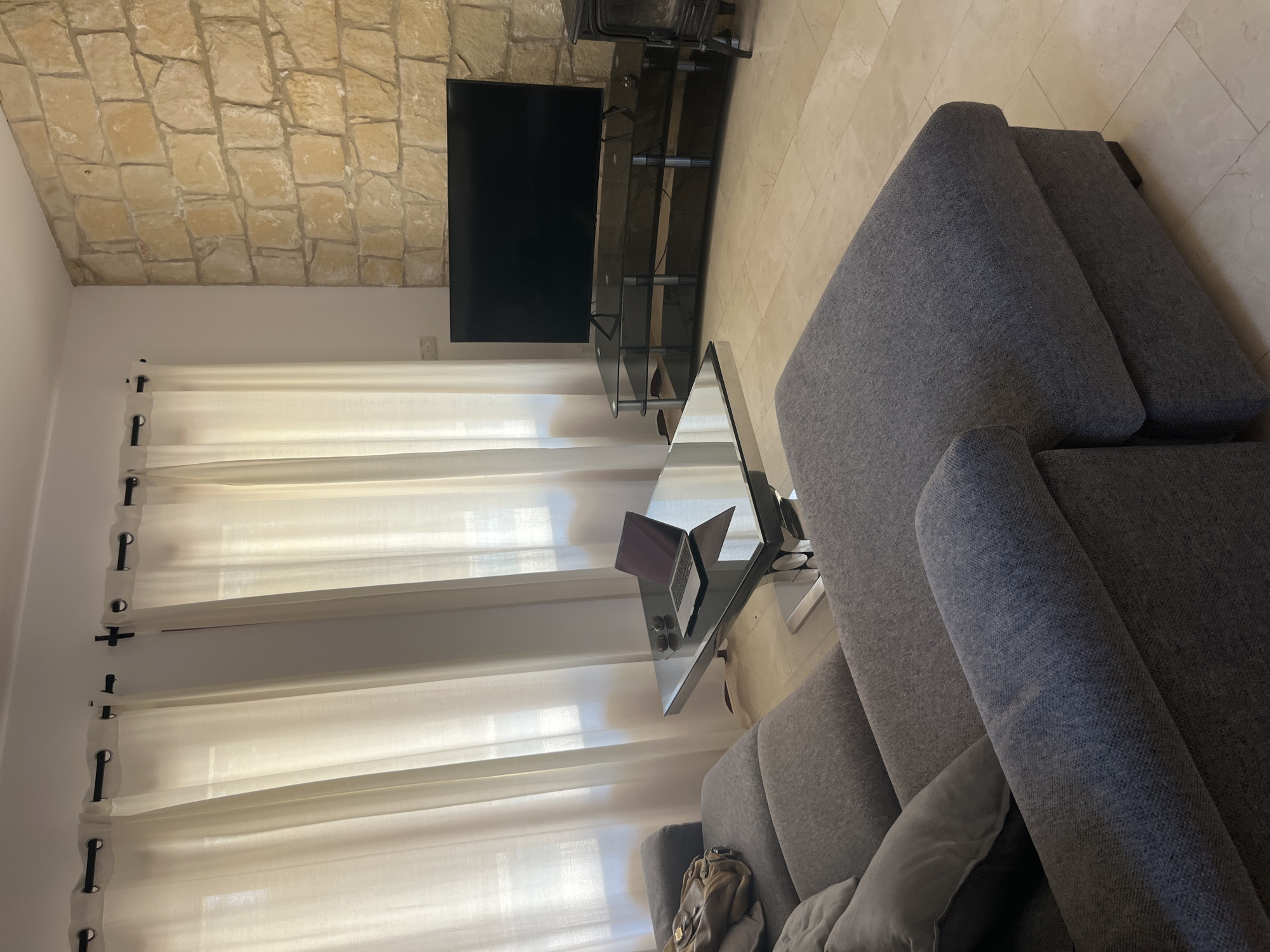 The image depicts a modern living room in Aphrodite Hills, Cyprus. A large, L-shaped grey sofa dominates the foreground, with cushions and a brown bag placed on it. In the center of the room, a glass coffee table with a chrome base holds a laptop and two sunglasses. Behind the sofa, sheer white curtains cover a window, allowing soft light to enter the room. To the right, a flat-screen television rests on a black glass entertainment unit against a textured stone wall. A dark wood-burning stove is partially visible in the far right corner. The floor is tiled with large, light-colored stone tiles. The overall impression is one of a comfortable and stylish living space.