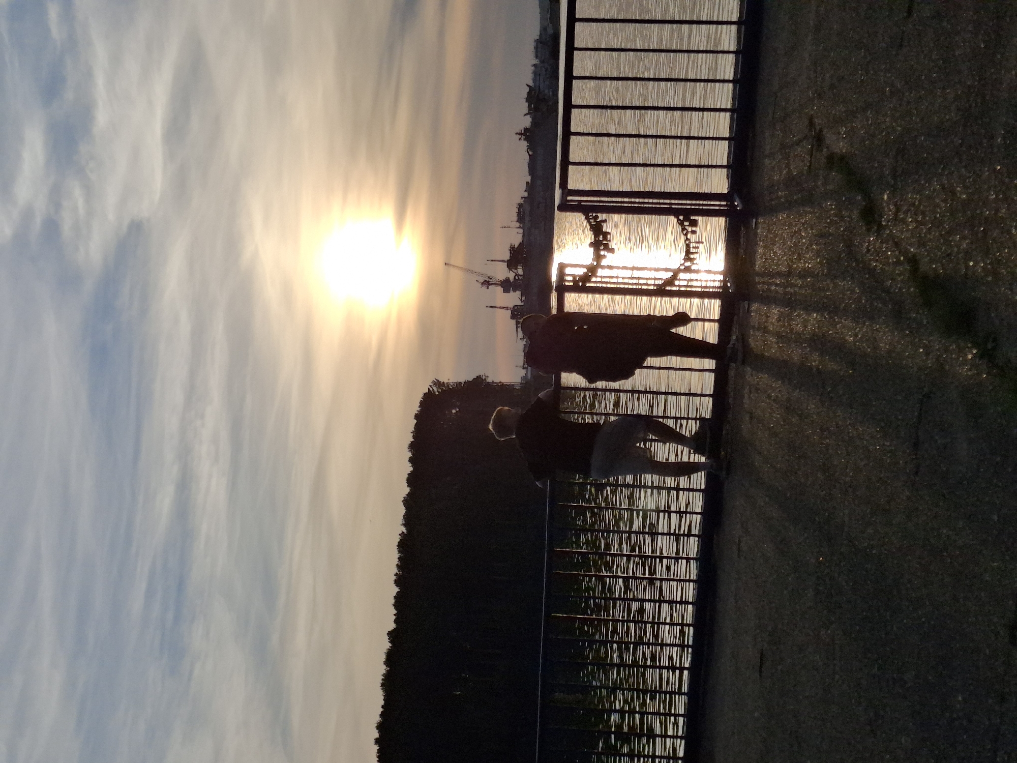 The image depicts a sunset scene on a body of water, likely the Kama River, adjacent to the city of Chaikovsky, Russia. Two individuals, silhouetted against the setting sun, stand on a paved walkway behind a black metal railing. The person on the left, appearing to be a male, has light-colored hair and is wearing shorts. They are leaning on the railing, facing the water. The person on the right is mostly obscured by shadow, but appears to be wearing a backpack. The sun, a bright orb, is positioned in the upper right quadrant of the sky, casting a warm glow and reflecting on the water's surface. Beyond the railing, a dense line of trees forms the background, and in the distance, industrial structures with cranes are visible. The sky is filled with wispy clouds, streaked with hues of orange and blue. Shadows from the railing and the figures are cast across the walkway.
