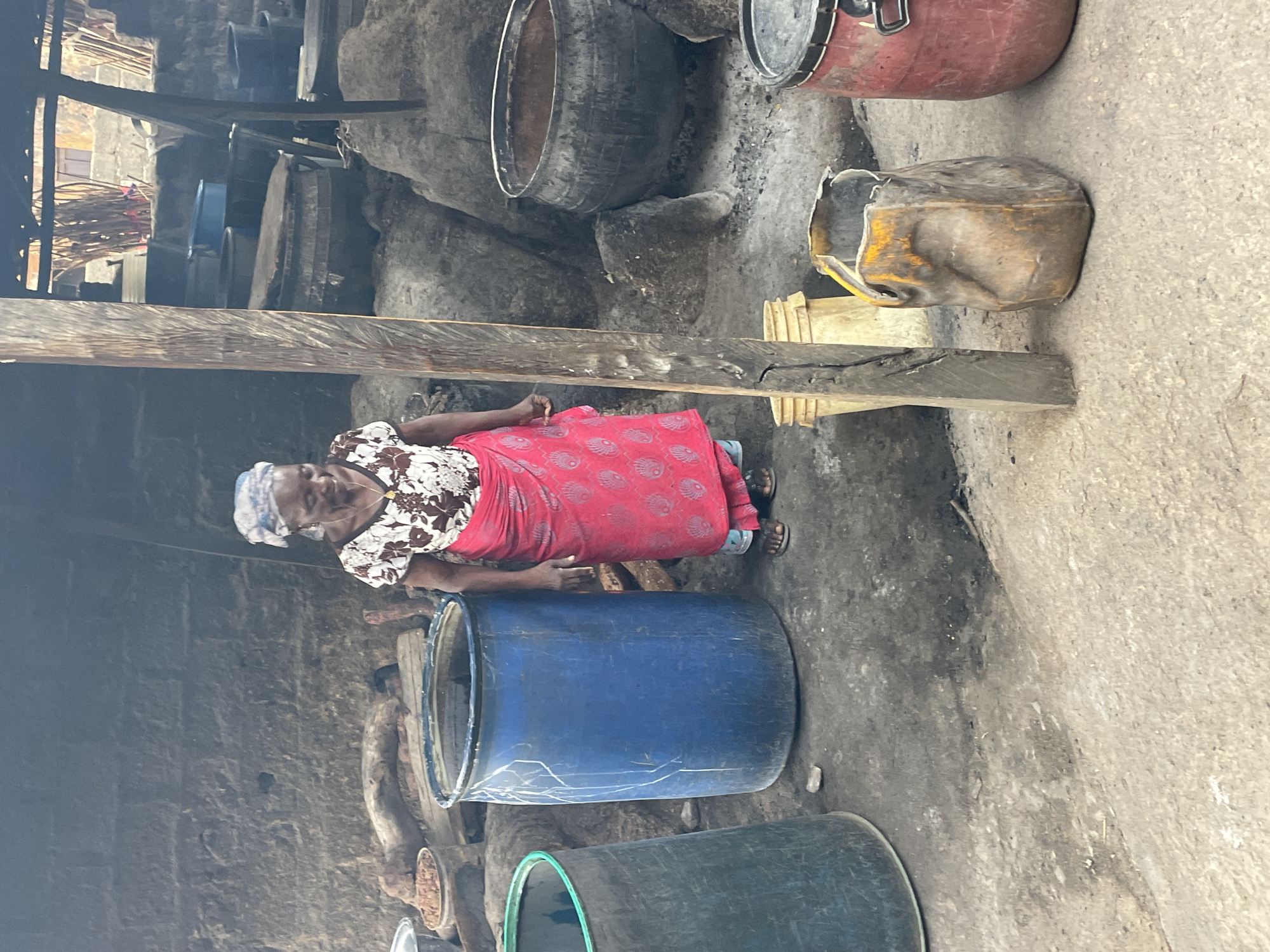 This image, taken in Jos, Nigeria, features an older woman standing in what appears to be an outdoor or semi-sheltered work and storage area. The woman is positioned towards the center-left, looking directly at the camera with a gentle smile. She is dressed in a white and brown patterned short-sleeved top, a long red skirt adorned with a repeating light-colored motif, a light blue and white patterned headscarf, a necklace with a pendant, and flip-flops.
The setting is characterized by an array of various large containers and barrels on a dirt or concrete ground. Prominently in the foreground, near the woman, is a large blue plastic barrel. Other visible containers include a dark green barrel, a red metal drum, and several dark grey/black barrels or pots of different sizes, some appearing stacked. A dented, yellow, open metal container rests on the ground to the right. A substantial wooden beam spans horizontally across the mid-ground, with other wooden structures visible in the background, suggesting a functional, perhaps makeshift, construction. Bundles of sticks or reeds are also noticeable in the upper left background.
The scene conveys an environment likely used for storage or a small-scale industrial process, typical of local production or material handling in the region. The natural lighting suggests it is daytime. The woman's calm expression and direct gaze create a sense of personal connection within the utilitarian setting. There is no discernible text visible in the image.