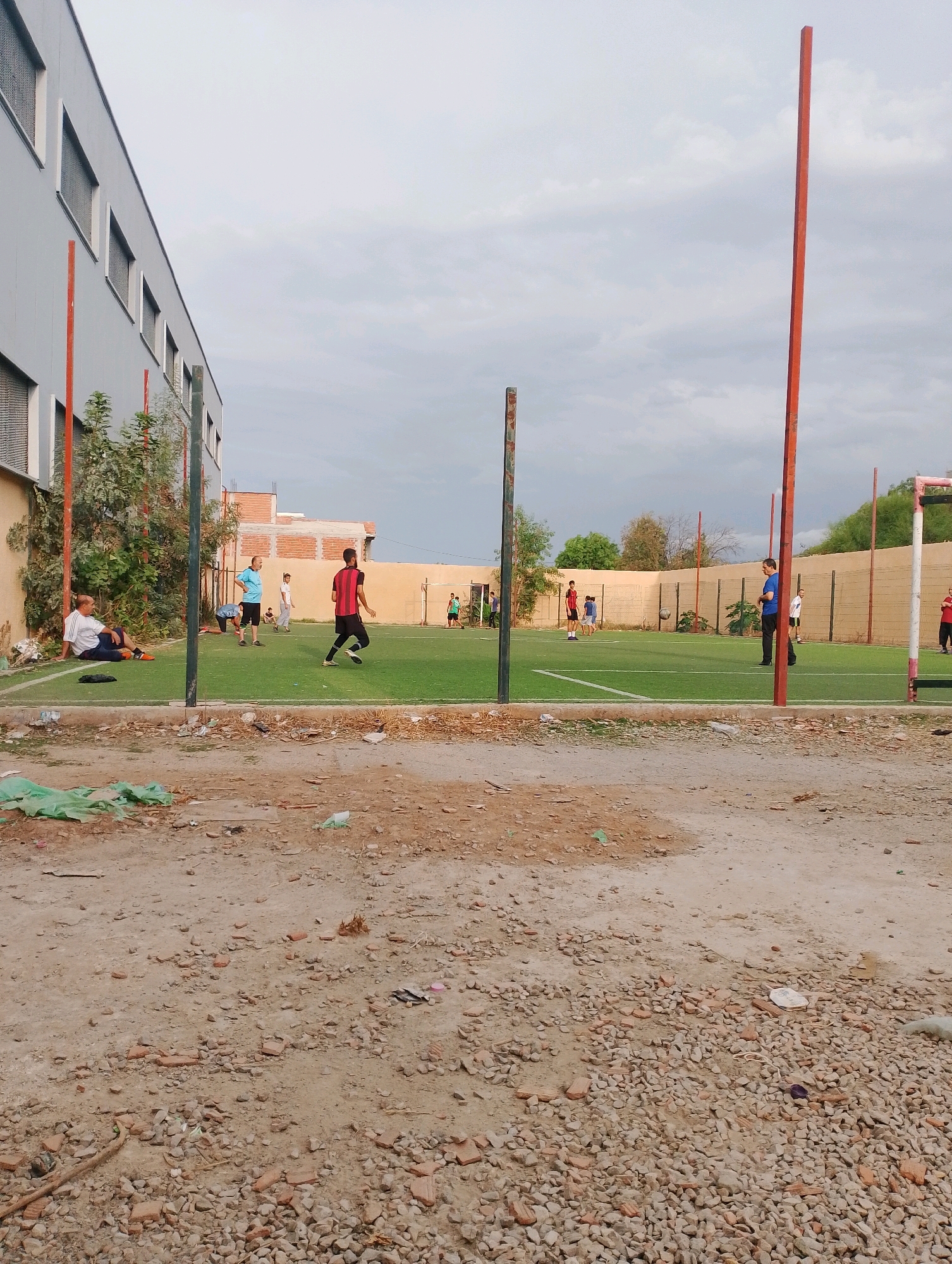 The image depicts an outdoor artificial turf soccer field in Bordj Menaiel, Algeria. The field is fenced and has soccer goals at either end. Several people are present on and around the field, some playing soccer, others watching or resting. The field is adjacent to a building and a dirt area covered with rubble and debris. The sky is overcast.