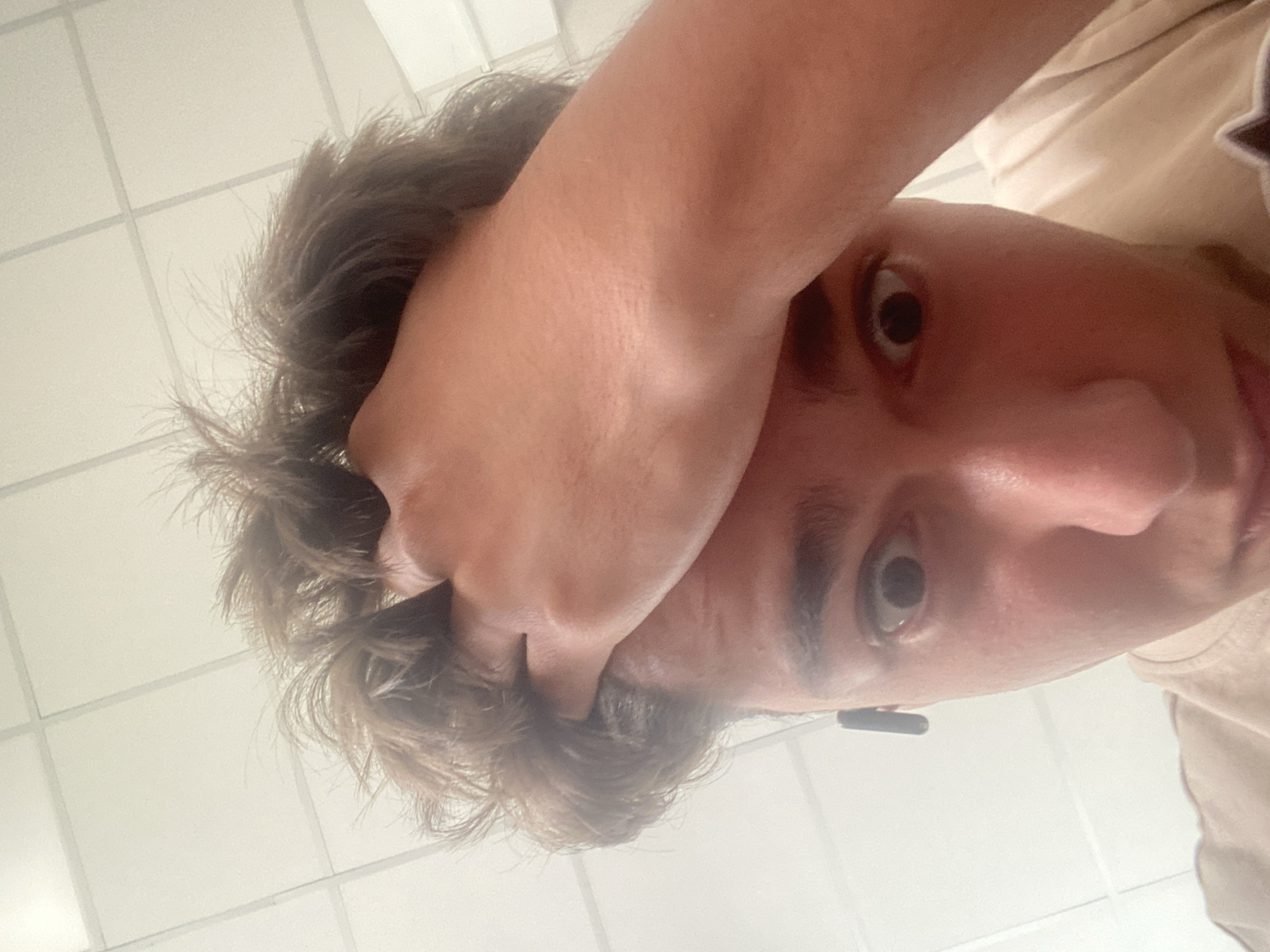 The image is a selfie taken from a low angle, looking up at a person. The person has light brown hair, fair skin, and dark eyes. One hand is raised and partially obscuring the forehead and hair. The person is wearing a light-colored shirt. The background appears to be a white tiled wall, possibly in a bathroom or similar space. There is no visible information to determine if the photo was taken in the city of Troyan, Bulgaria.