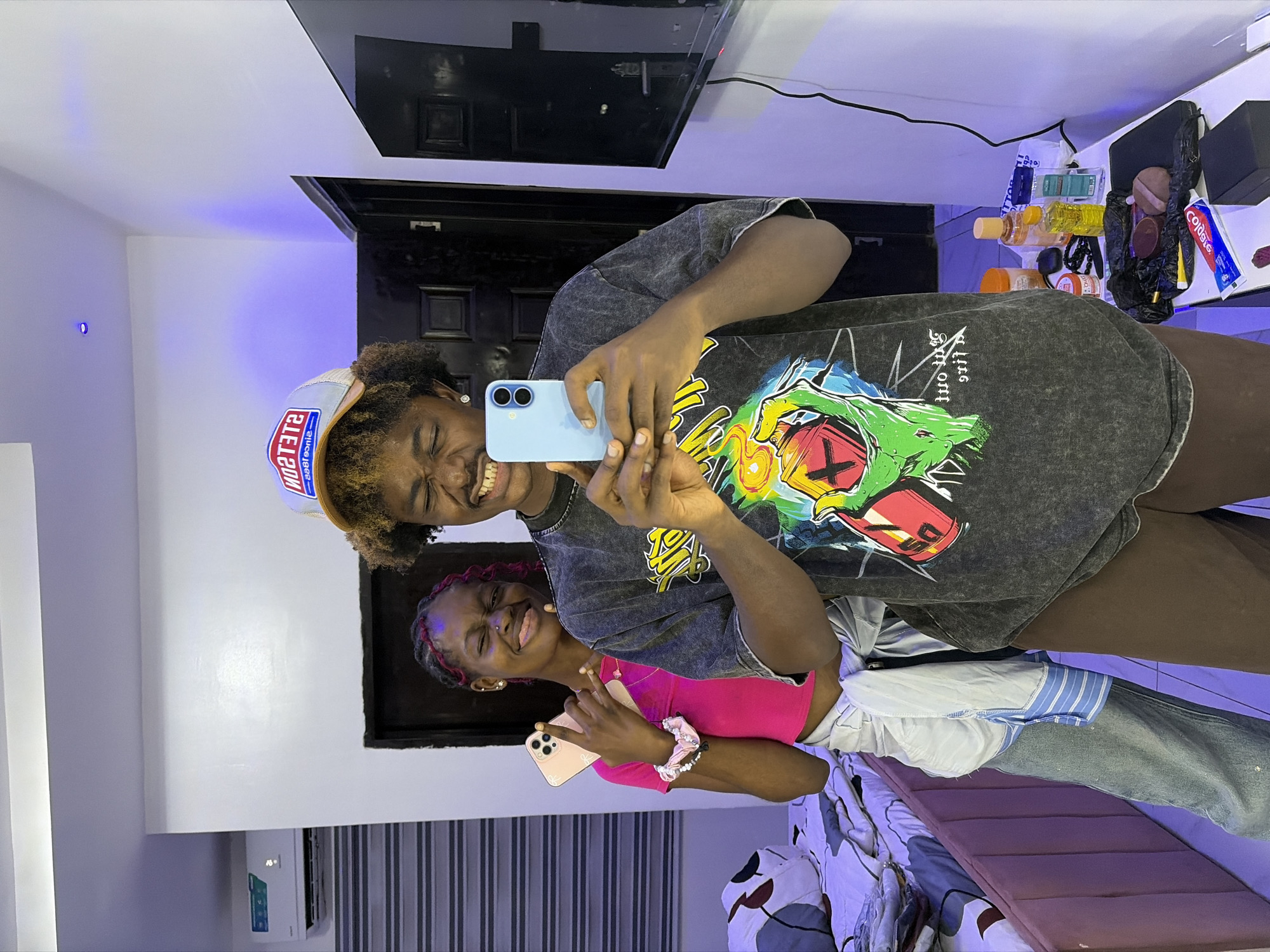 Two young women are captured taking a mirror selfie indoors in Igboefon, Nigeria. Both subjects display wide, joyful smiles, conveying a happy and playful mood. The image is rotated 90 degrees clockwise, indicating it was taken in portrait orientation and is presented sideways.
The primary subject, positioned closer to the mirror, has light brown curly hair and wears a light-colored baseball cap with the reversed text "NOS TET2" and "5681 ecnis" (likely "STETSON since 1865"). She is dressed in a dark gray oversized t-shirt adorned with a large, colorful graphic design, including some reversed text that appears to be "THE OFF". She holds a light blue smartphone with both hands, using it to capture the reflection, and her eyes are nearly closed in a wide smile.
Behind her, the second subject has dark braided hair with pink highlights. She wears a bright pink crop top, and a light blue and white striped shirt is tied around her waist, paired with light blue jeans. She holds a light pink smartphone in one hand and makes a peace sign with the other, also smiling broadly at the camera. A pink scrunchie is visible on her wrist.
The indoor setting features white walls, with a subtle blue/purple LED light visible in the upper left, suggesting a controlled ambiance or evening time. In the background, visible both directly and in the mirror reflection, are a large flat-screen television, a black door or cabinet, and a white desk or shelf to the right. This desk holds various items, including yellow and orange bottles, a black bag, and a tube of "Colgate" toothpaste. An air conditioner unit is mounted on the wall to the left, above a wall with vertical grey and white stripes. Part of a bed with a patterned duvet is visible at the bottom right of the frame.