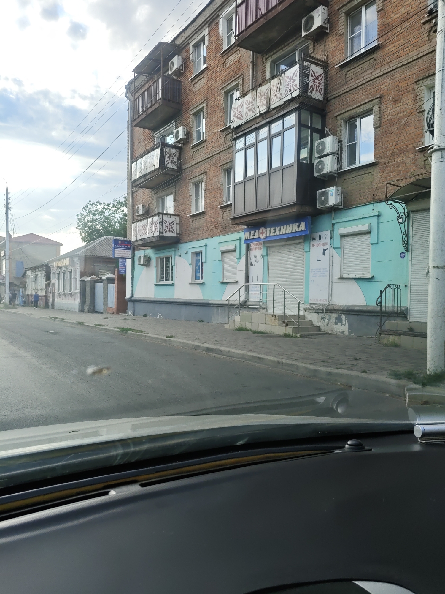 A storefront with a bright turquoise and white facade is visible on a brick building in Novocherkassk, Russia. The storefront has roller shutters over its windows and doors, and a blue sign above the entrance reads "МЕДТЕХНИКА," which translates to "Medical Equipment." On the facade, there are images depicting medical devices and orthopedics. The building has several balconies with ornate railings, and air conditioning units are mounted on the exterior walls. A sidewalk and a street with a few scattered leaves are in the foreground. The sky above is partly cloudy.