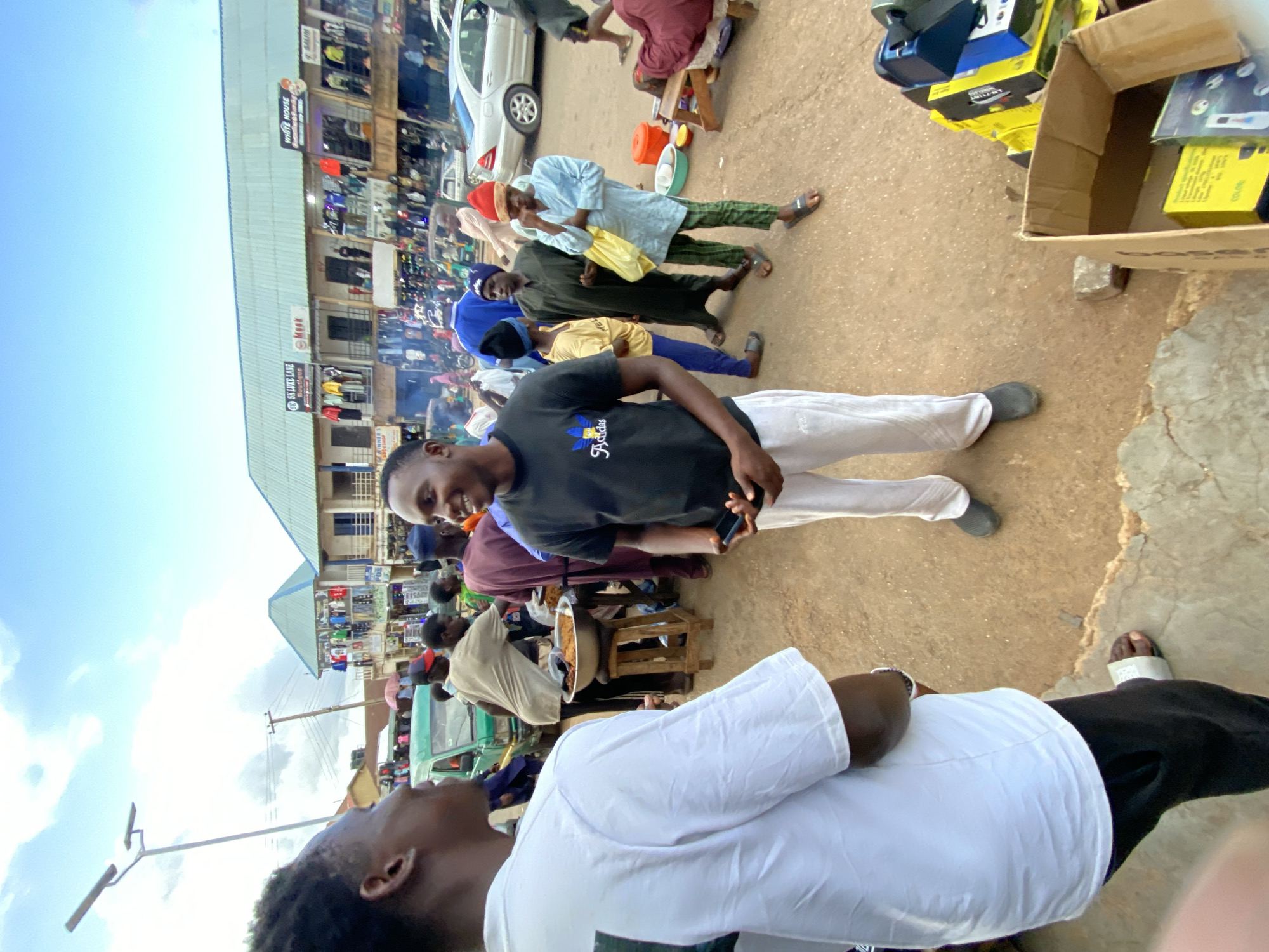 The image captures a bustling street scene in Bukuru, Nigeria. The area appears to be a commercial district with multi-story buildings housing shops and businesses visible in the background. People are seen walking, some carrying items, suggesting a market or shopping area. The ground is a mix of dirt and concrete.
The main subjects are two men. One, wearing a black t-shirt and white pants, is looking directly at the camera while holding a mobile phone. Another man, closer to the viewer, is wearing a white shirt and has his back to the camera.
Other visible elements include:
* A car parked nearby.
* Boxes containing what appear to be electronic goods.
* Street vendors selling food from bowls.
* Other pedestrians of varying ages and clothing styles.
* Overhead electrical wires.
* Shop signage on the buildings.