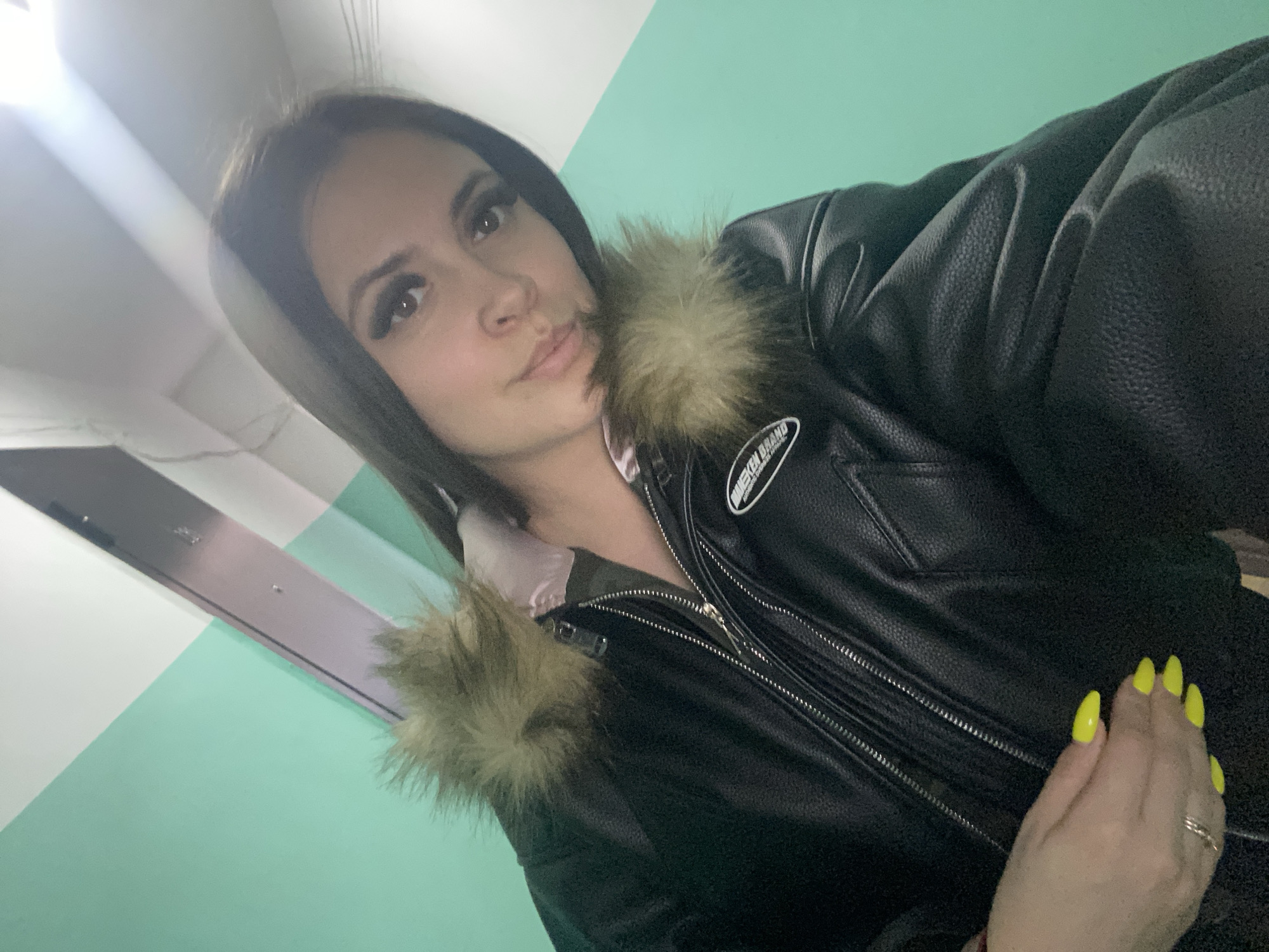 A woman is pictured indoors. She has long, dark hair and is wearing a black leather jacket with fur trim around the hood. She has yellow painted fingernails. The background consists of a pastel green and white wall. The location is the city of Chaikovsky, Russia.