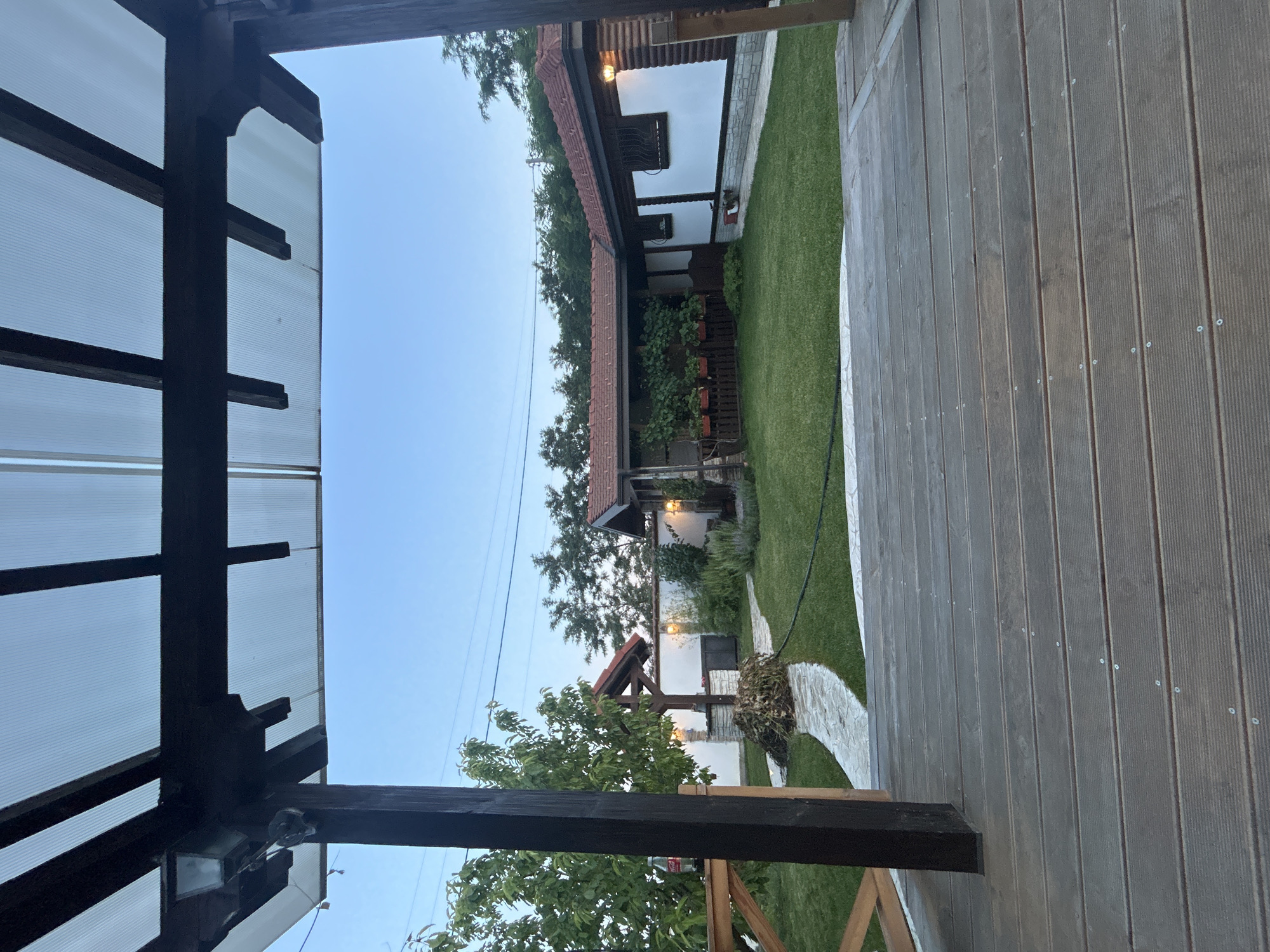 The image captures a view of a backyard in Voyvodino, Bulgaria, from a wooden deck. The foreground is dominated by the wooden planks of the deck, with a dark wooden support post on the left. Beyond the deck, a well-maintained lawn stretches out towards a house with a red-tiled roof. To the left of the lawn, a stone pathway leads to a rustic well with a red roof. The house, partially visible on the right, has white walls with stone detailing at the base and wooden accents. Several outdoor lights are visible, some illuminating the pathway and the house exterior. Trees and foliage surround the property, and the sky is clear and light blue, suggesting it is either early morning or late evening. There are no people or animals visible in the image.