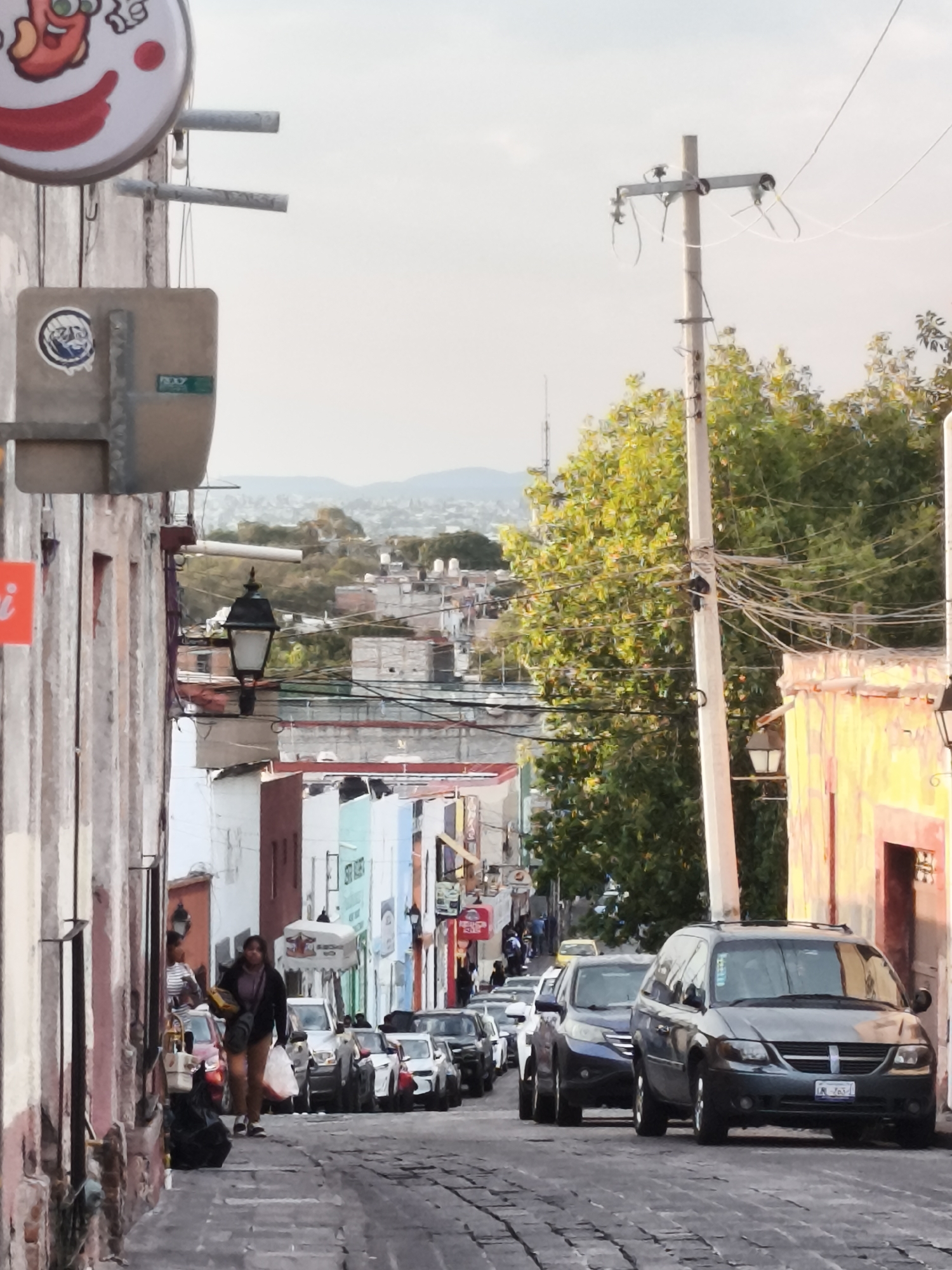 The image depicts a narrow, cobblestone street in Querétaro, Mexico, with buildings of various colors and heights lining both sides. The street slopes downward and is filled with parked cars, including a grey minivan prominently positioned in the foreground. A utility pole with numerous electrical wires stands to the right of the street, adjacent to a building with a yellow-painted facade. A woman carrying bags walks along the left side of the street. In the background, there are rooftops and distant hills visible under a hazy sky. Signs and other urban fixtures are attached to the buildings, contributing to the overall atmosphere.