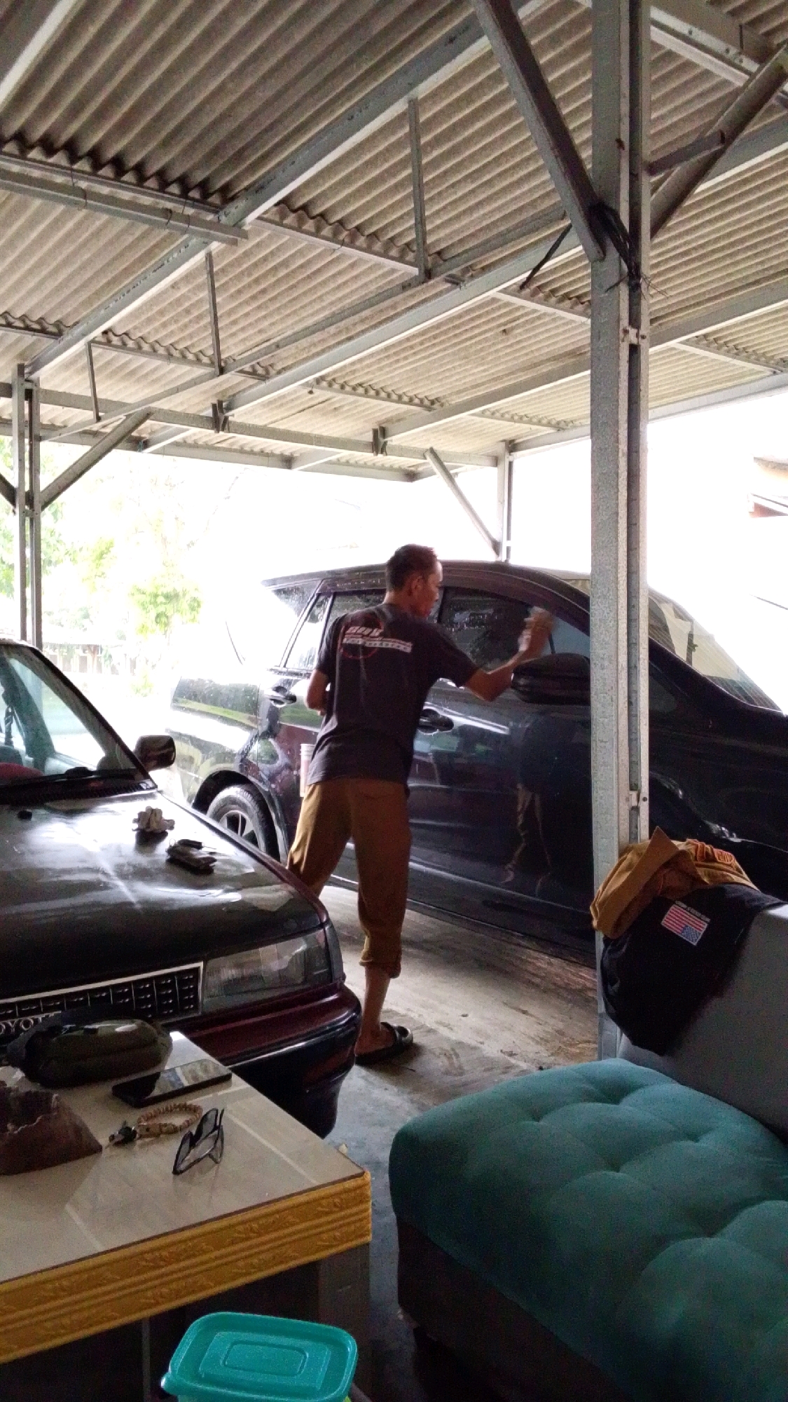 In Cintaratu, Indonesia, a man is captured mid-action washing a dark-colored car. He is wearing a dark t-shirt and brown trousers, and is seen using a sponge to clean the vehicle's side. The scene appears to be under a corrugated metal roof supported by metal beams, suggesting it might be a carport or an open-sided garage. In the foreground, the hood of a red car is visible, with a table to its left displaying various items including a green pouch, a mobile phone, a bracelet, and a pair of glasses. A turquoise plastic container rests on the floor in front of the table. A large, dark-colored SUV is parked behind the car being washed. The environment appears to be outdoors, with natural light filtering through the roof structure.