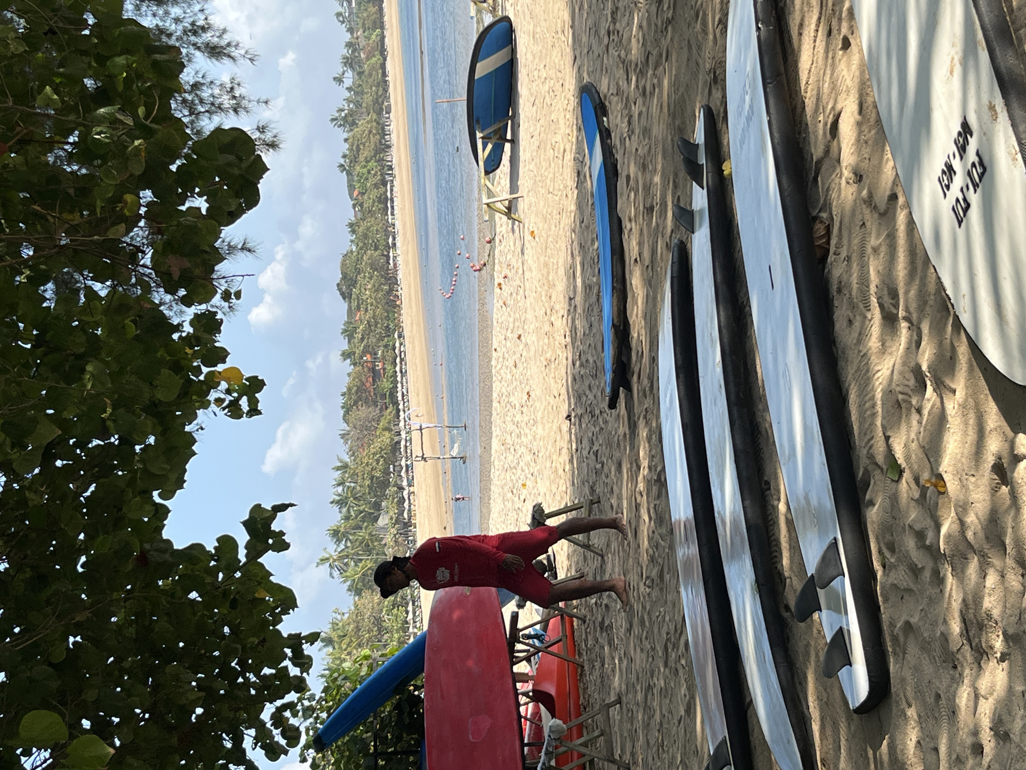 In Kuta Selatan, Indonesia, on a sandy beach under a canopy of green foliage and blue sky, a man in a red long-sleeved shirt and red shorts walks barefoot across the sand. He is near several surfboards, one large red surfboard propped up with a blue surfboard on top, and other smaller red and orange objects. In the background, the ocean stretches towards a distant tree-lined coast with buildings and white umbrellas. Several surfboards are arranged on the sand in the foreground. The scene is bright and sunny, with clear water and a few buoys marking an area in the ocean.