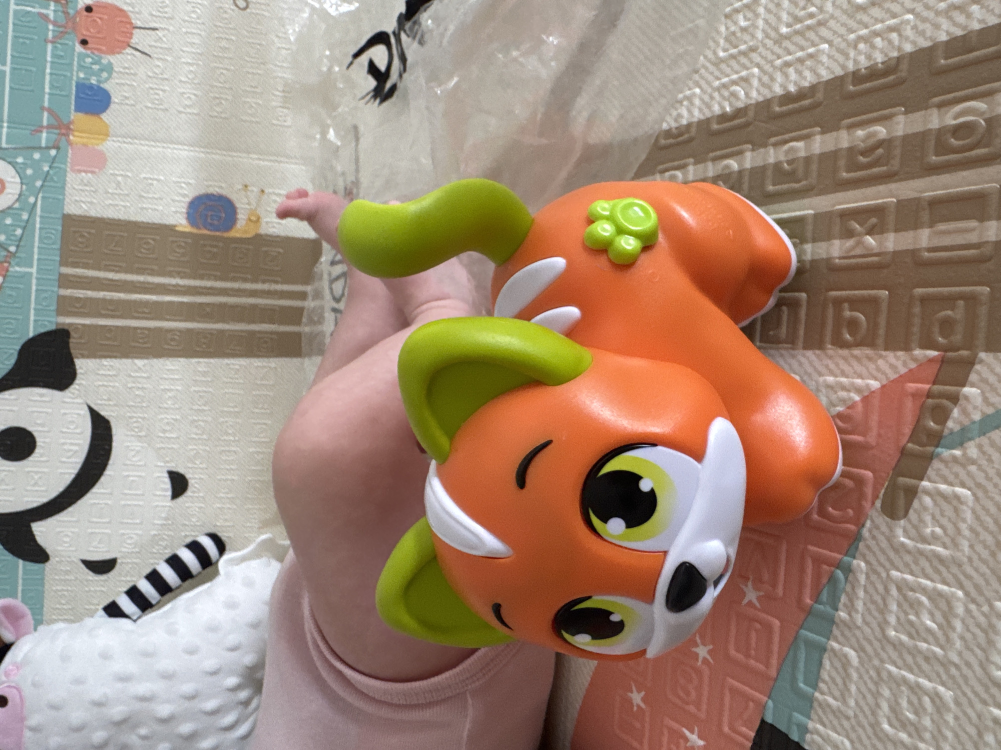 An overhead medium shot captures a portion of a baby lying on a colorful playmat. The baby is wearing a pink onesie and their legs are visible. To the right of the baby's legs, a bright orange plastic toy animal resembling a fox or dog is positioned on the playmat. The toy has large, round green and yellow eyes, green ears, and a white muzzle. A small green paw print design is visible on its side. The playmat beneath the subjects features a grid pattern with various numbers and symbols embossed into its surface. In the background, a white plush toy with black and white stripes is partially visible. The overall scene appears to be indoors, on a playmat, with no discernible elements indicating a specific location in Isola delle Femmine, Italy.