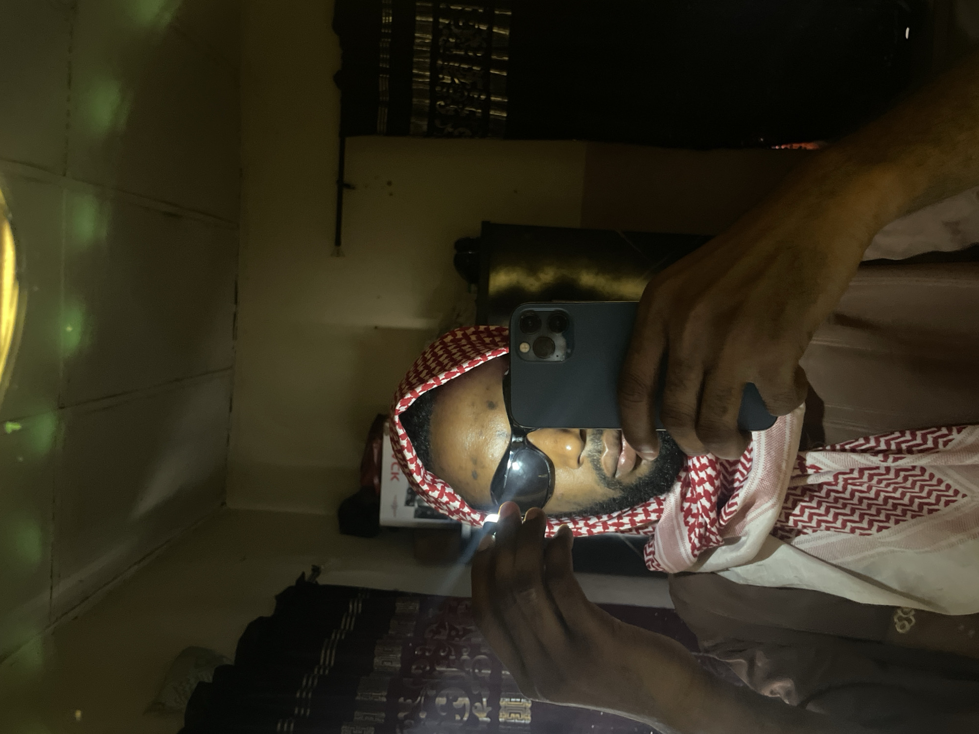 A man wearing a red and white patterned headscarf and sunglasses is taking a selfie in a mirror with a black phone. The environment appears to be a room. The location is unknown, and there is no evidence to suggest it is in Lokoja, Nigeria.