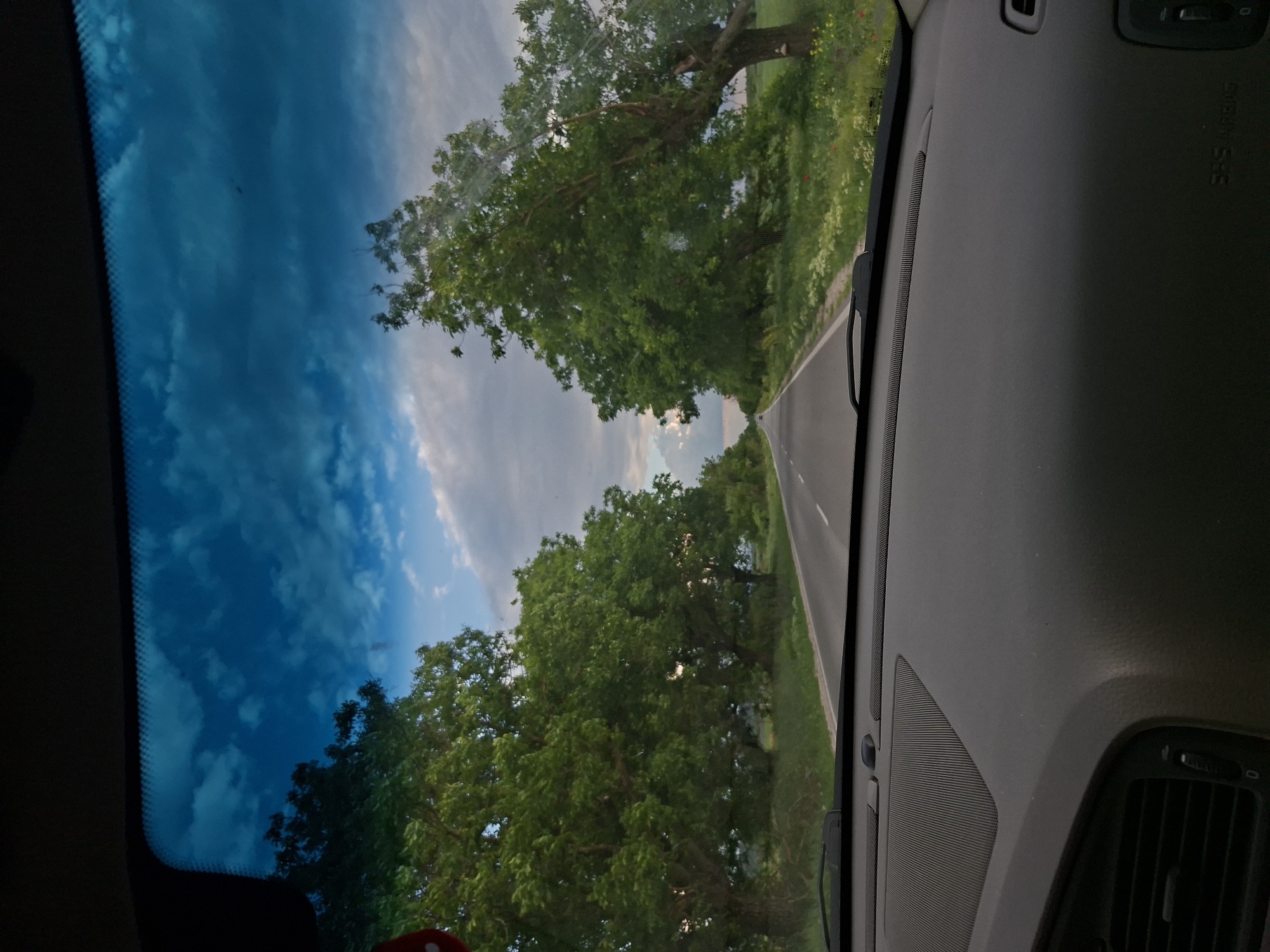The image shows a view from inside a car, looking out through the windshield. A narrow asphalt road stretches ahead, flanked by lush green trees on both sides, forming a canopy above the road. The sky is visible through gaps in the trees, displaying a mix of blue sky and white clouds. The car's dashboard is visible in the foreground, with features like vents and airbags clearly discernible. Based on the road type and vegetation, the location is likely a rural area. I do not see specific clues allowing me to place this view as in the city of Pobeda, Bulgaria.