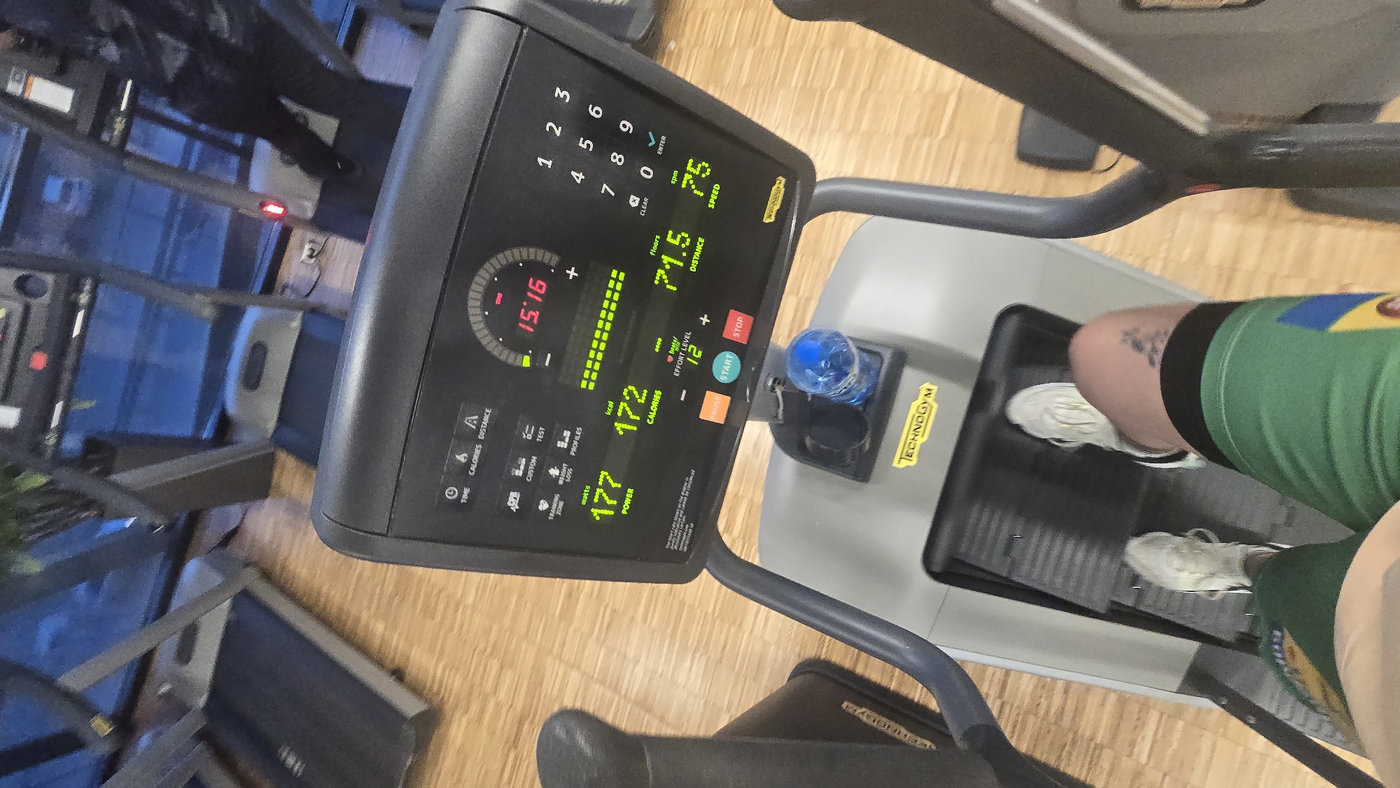 This high-angle, slightly rotated image captures a person exercising on a Technogym elliptical machine in a gym located in Sofia, Bulgaria. The perspective looks down towards the machine's console and the person's legs.
The main subject is the fitness equipment, specifically the console of an elliptical machine, displaying various workout statistics. The console shows a time of "15:16", "71.5" floors, "75" strokes per minute (spm), "172" calories burned, "177" watts of power, and an "EFFORT LEVEL" of "12". A numeric keypad (1-9, 0, CLEAR, ENTER) is also visible. Other labels on the console include "TIME," "CALORIES," "DISTANCE," "CUSTOM," "TEST," "PROFILES," "START," and "STOP." The brand "TECHNOGYM" is prominently displayed on the console and the machine's base.
A blue water bottle is in the machine's holder. Below the console, the legs of the person exercising are visible, wearing green and yellow athletic shorts, white sneakers, and black socks. A small tattoo is visible on the upper left thigh. The flooring is light wood. In the background, other gym equipment, resembling treadmills, can be seen, along with reflections that suggest large windows. The overall setting is a modern fitness facility.