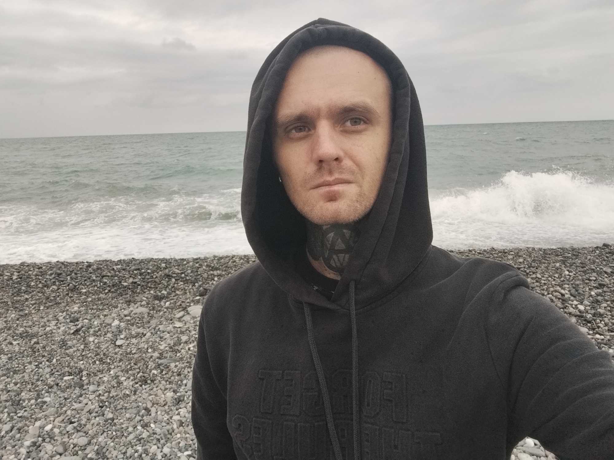 A man wearing a dark grey hooded sweatshirt and a dark t-shirt underneath is taking a selfie on a pebble beach with the sea in the background. The sky is overcast. The man has light skin, short dark hair, and a beard. He has a tattoo on his neck. The sweatshirt has embossed lettering on the front. The sea is rough with waves crashing on the shore. The pebble beach is composed of grey, white, and black stones. The scene is likely in Batumi, Georgia, as it depicts a typical Black Sea coastline.