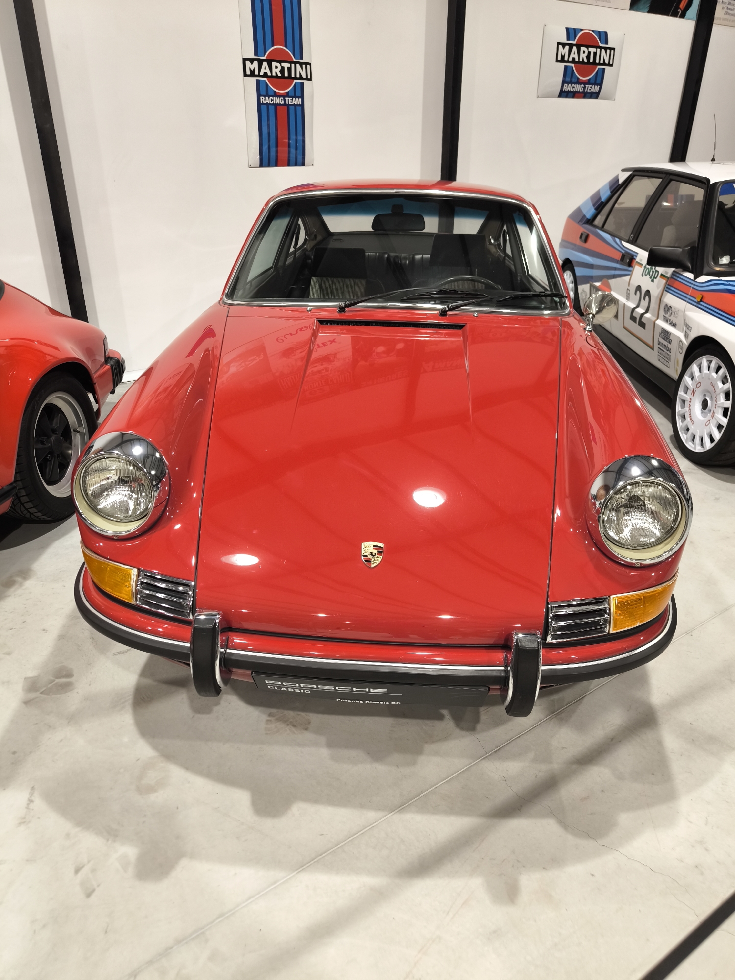 The image shows the front of a red vintage Porsche car in a showroom. The car is well-lit and positioned centrally in the frame. Behind the Porsche, there's a white car with "22" and racing decals, and two "Martini Racing Team" banners are visible on the wall. The showroom has a white floor and walls, with black vertical stripes visible in the background. The Porsche emblem is visible on the hood. The location, Kapatovo, Bulgaria is not discernible from the visual elements in the image.