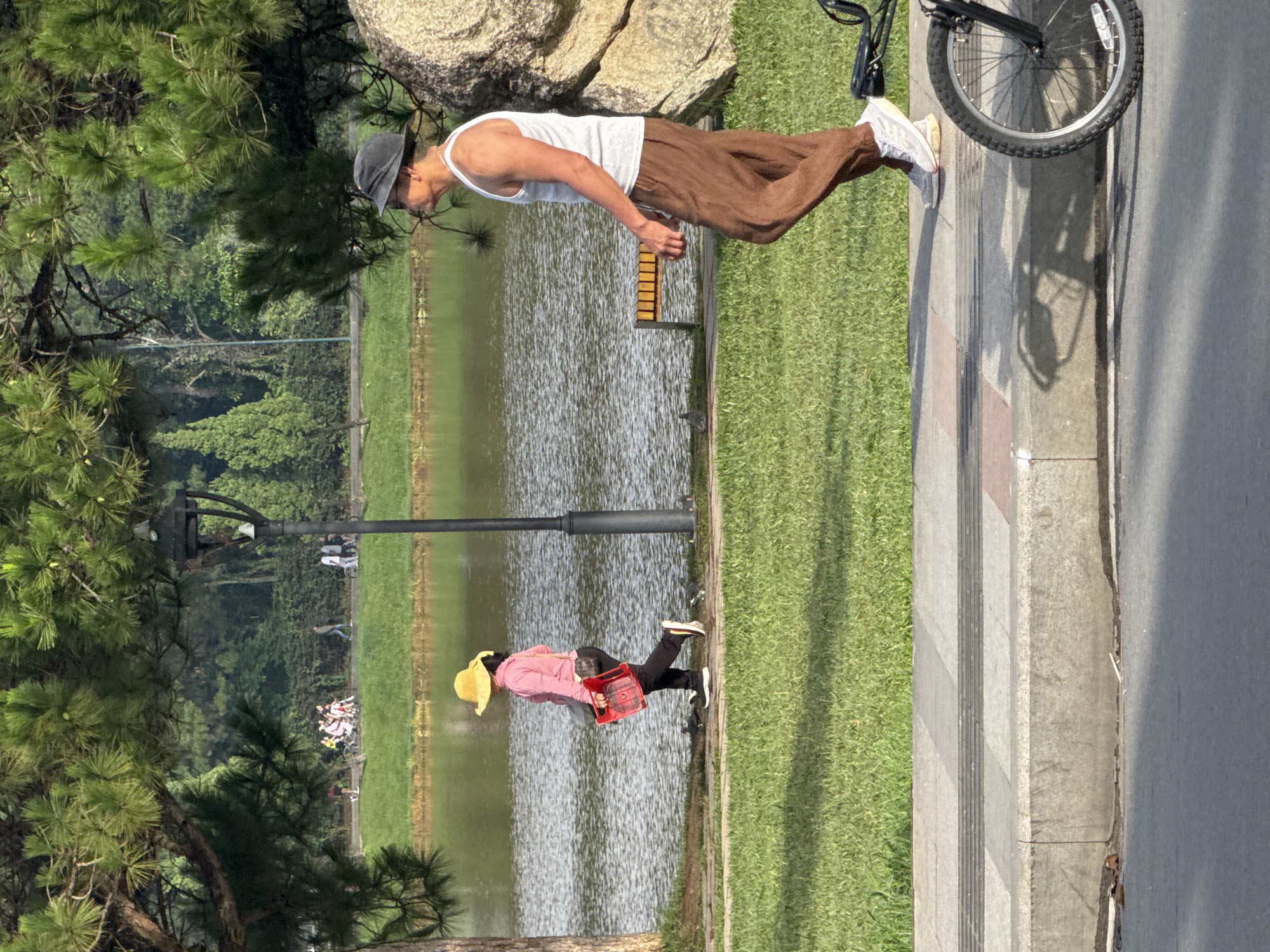 This image captures a scene at Xuan Huong Lake in Da Lat, Vietnam. In the foreground, a man wearing a white tank top, brown pants, a grey hat, and white sneakers walks along a paved path. To his left, a woman in a pink jacket and yellow hat, carrying a red basket, walks in the opposite direction. Behind them, a body of water reflects the surrounding trees and sky. Further in the background, a group of people are seen near trees and a pathway, with some appearing to be on bicycles. The environment is characterized by lush green grass, mature trees, and a lamppost. The lighting suggests it is daytime, with shadows cast on the path and ground. A bicycle wheel is partially visible in the bottom right corner of the frame.