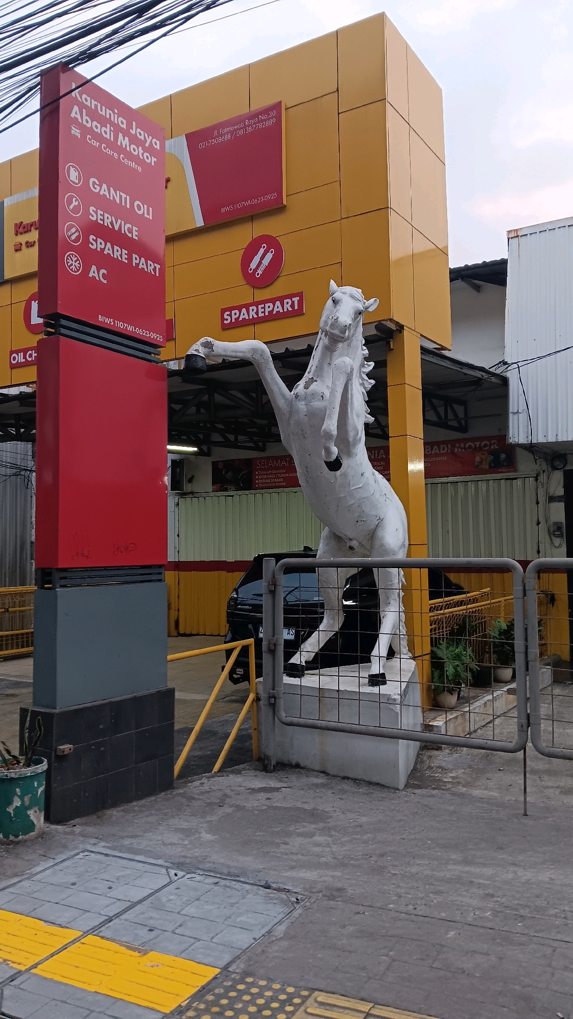 An outdoor shot captures the facade of a car care center in South Jakarta, Indonesia. The building features a bright yellow exterior with prominent red signage. To the left, a tall red signpost lists services offered, including "GANTI OLI," "SERVICE," "SPARE PART," and "AC." In front of the building, a white statue of a horse rears up on a concrete pedestal. The ground in front of the statue is paved with concrete and includes tactile paving strips. A dark-colored car is parked behind the horse statue, partially obscured. The scene is framed by metal fencing and yellow poles, suggesting a service or entrance area.