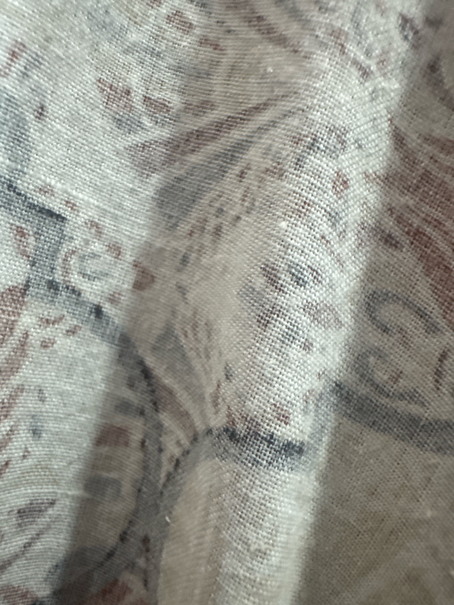 The image is a close-up, textured photograph of a woven fabric with a paisley-like pattern. The fabric appears to be a light, neutral color, possibly linen or a linen blend, with a visible weave. The pattern consists of blurred, organic shapes in muted tones of brown, grey, and possibly a dark blue or black. The fabric is creased and folded, creating shadows and highlights that emphasize its texture and dimension. The overall impression is one of a decorative textile. The image does not contain any discernible elements that would allow for its localization to Rufisque, Senegal, or any other specific geographical location.