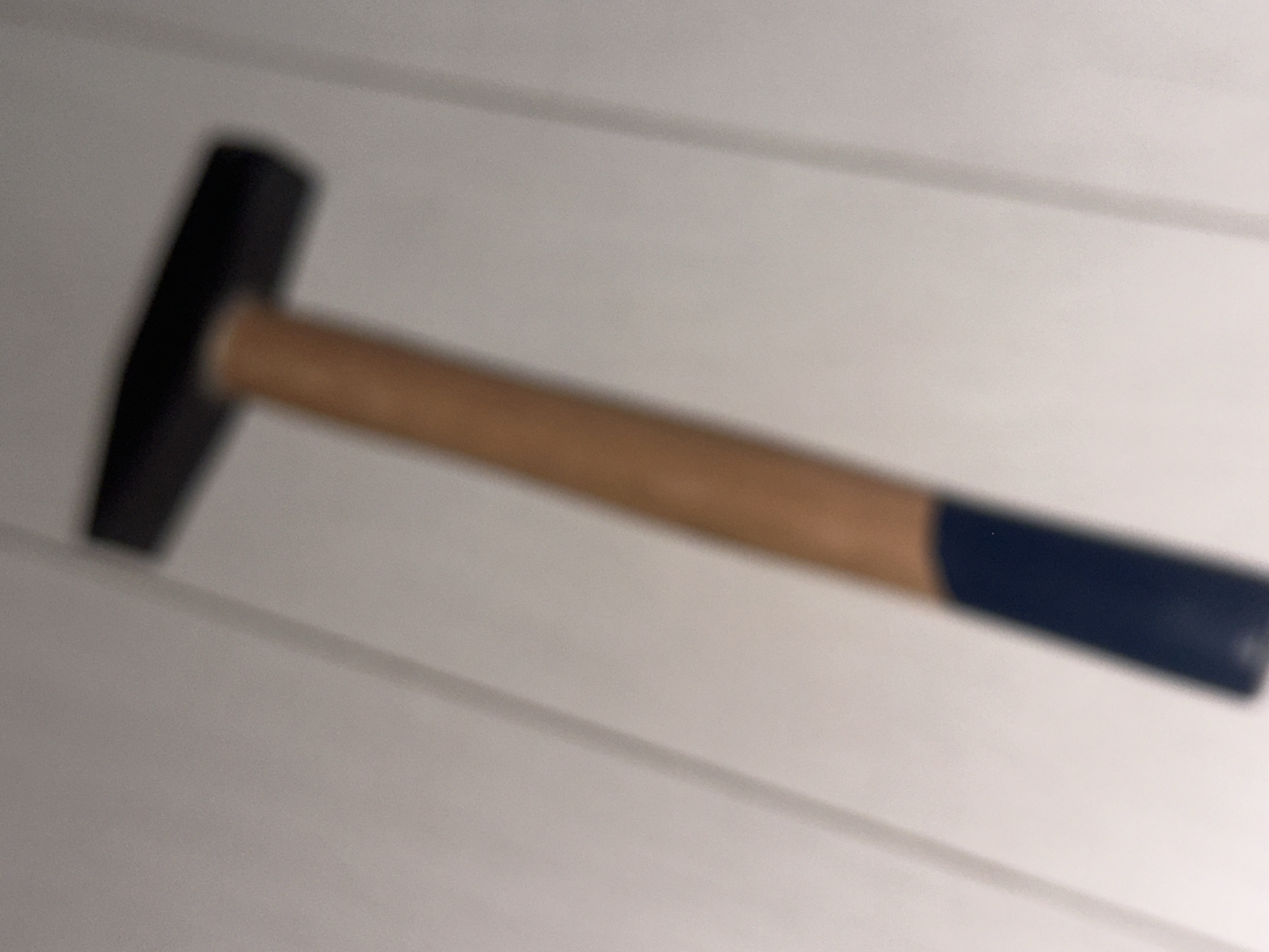 A hammer with a black head and a wooden handle with a dark blue grip is positioned diagonally on a light-colored surface, likely a floor or wall, with faint lines suggesting a wood-paneled or planked texture. The hammer is the sole object in focus. The scene appears to be indoors, and no specific location in Sofia, Bulgaria can be determined from the provided image. The image quality is low, with significant blur.