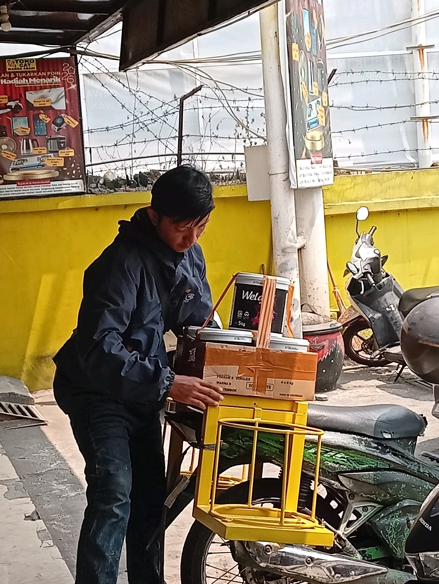 A man in a dark blue jacket and jeans is standing next to a yellow metal rack attached to a motorcycle. He is holding two containers, one a tall, white cylinder with black labeling, and the other a rectangular cardboard box also with labels. The man appears to be placing these items onto the rack. The motorcycle is parked on a paved surface with a yellow wall in the background. A poster is attached to the wall to the left, and another banner hangs from a pole on the right. There are barbed wire fences visible above the wall. The scene appears to be outdoors in a commercial or industrial area.