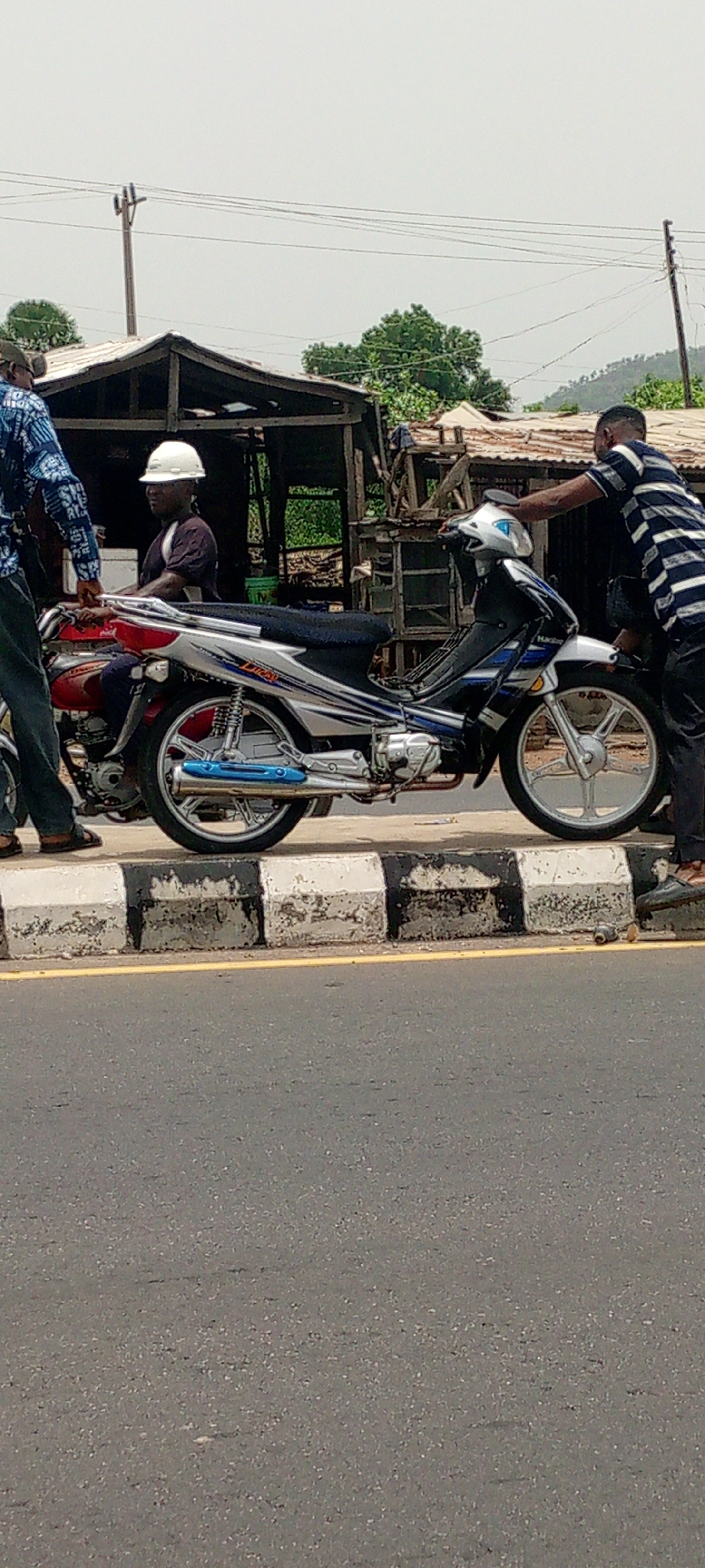 The image shows two motorcycles parked near a road in what appears to be Zango-ndaji, Nigeria. One is red and the other is silver and blue. A man in a white helmet is seated on the red motorcycle. Another man wearing a striped shirt is touching the silver and blue motorcycle. A third man, wearing a blue and black patterned shirt, stands near the motorcycles. A building, possibly a shop, is visible behind the motorcycles. A road with a yellow line and a black and white curb is in the foreground. Hills are visible in the background.