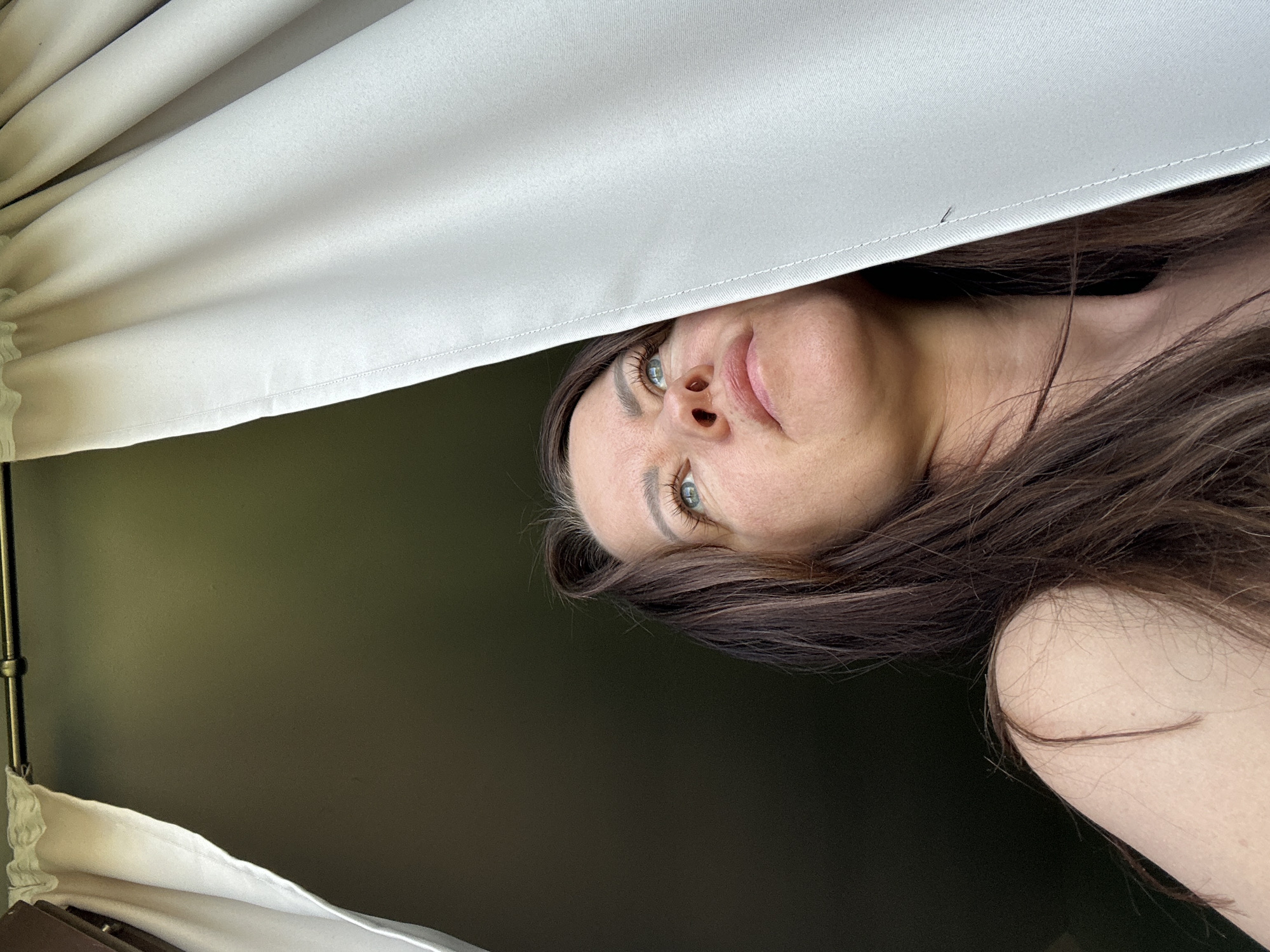 A woman with long brown hair and blue eyes looks down at the camera from under a white curtain. The background is a solid dark green. The location is unknown, though the prompt specifies the city of Chaikovsky, Russia. The image focuses on the woman's face, and her expression is neutral. Her right shoulder is visible in the lower right corner. The curtain is gathered on a metal rod.