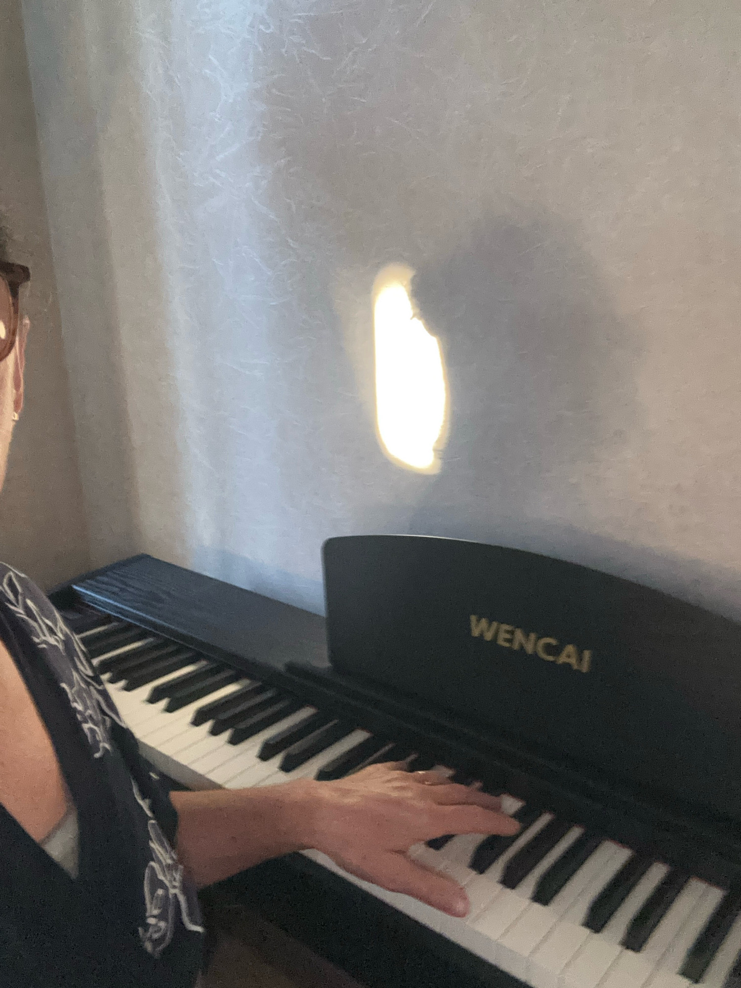 A person with brown-rimmed glasses, wearing a dark blue patterned shirt, is playing a WENCAI digital piano. The piano is black with white and black keys. A light source is casting a bright oval-shaped reflection on the textured, light-colored wall behind the piano. The scene appears to be indoors, in a room with soft lighting, likely during the day.