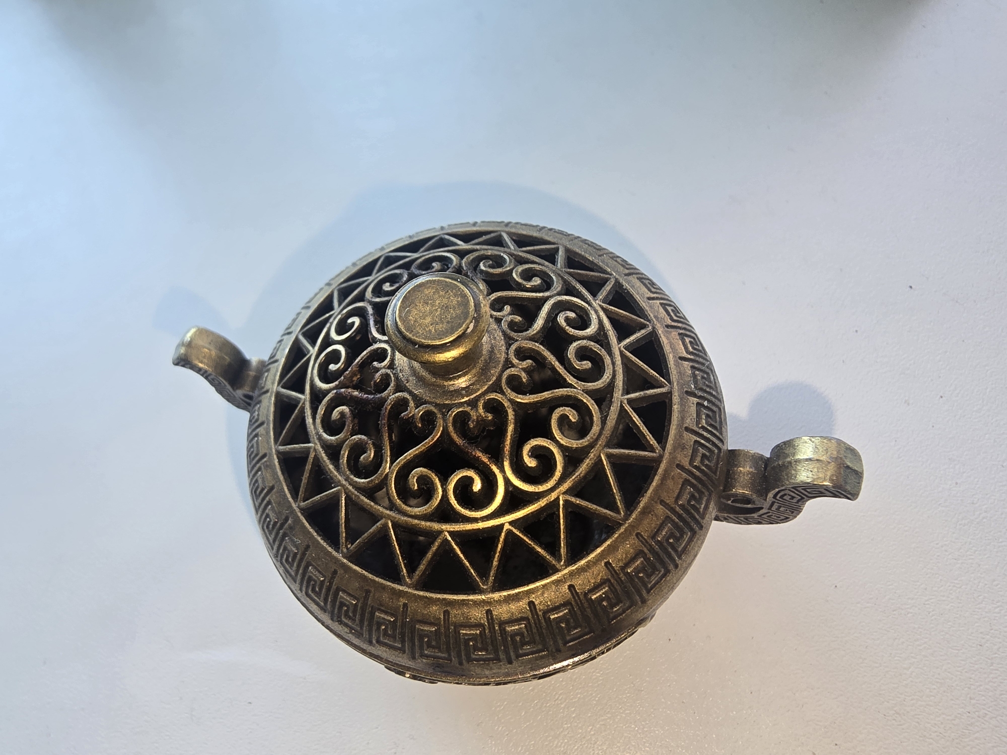 A circular, metallic object with intricate patterns rests on a white surface. The object has a decorative lid with ornate scrollwork and a Greek key border. Two small, curved handles extend from the sides. The lighting suggests a studio setting or controlled indoor environment, casting a shadow to the right of the object. There is no indication of a specific city or geographical location.