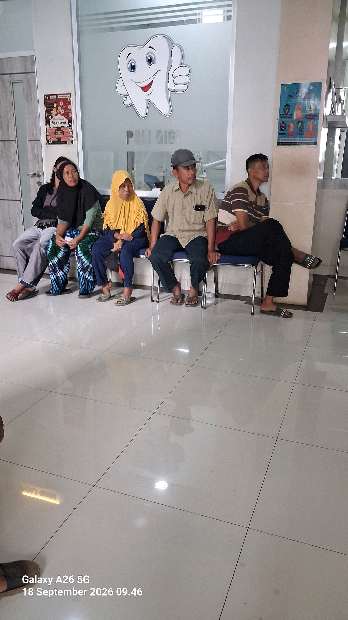 In a waiting area, likely a dental clinic, four individuals are seated on a bench. To the left, a woman wearing a black hijab and a patterned skirt sits beside another woman also in a hijab, this one yellow, with a blue garment. To their right are two men; the first in a light-colored collared shirt and dark pants, and the second in a striped polo shirt and dark pants. A large, cartoonish tooth graphic with a thumbs-up is displayed on the frosted glass door behind them, with "POLI GIGI" written below it, indicating a dental service. Informational posters are visible on the wall to the left and right, one depicting health-related icons and text. The floor is tiled and reflective. The timestamp "Galaxy A26 5G 18 September 2026 09.46" is embedded in the lower-left corner of the image.
