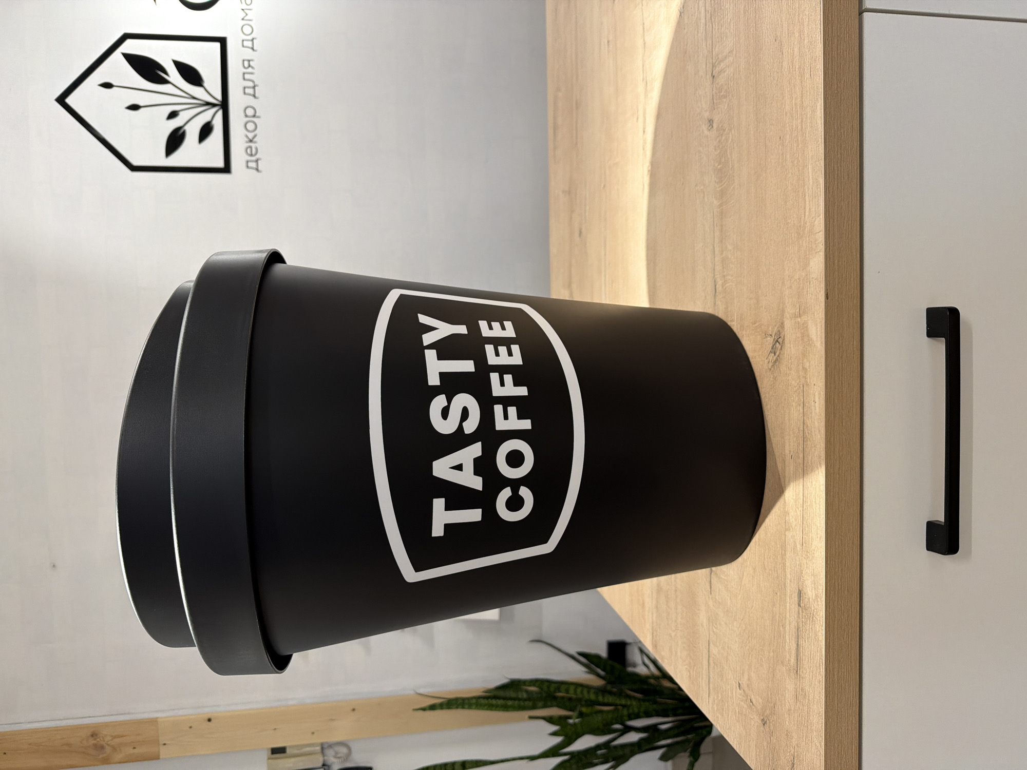 A large, black cylindrical coffee cup with a lid is prominently displayed, standing upright on a light-colored wooden surface. The cup features a white logo that reads "TASTY COFFEE" enclosed within a hexagonal or shield-like badge.
Behind the wooden surface, a plain white wall serves as the backdrop. Mounted on the wall is a black decorative element shaped like a stylized house, from which leafy branches extend. Adjacent to this logo, text in Cyrillic is visible, spelling out "ДЕКОР ДЛЯ ДОМА", which translates to "DECOR FOR HOME".
To the right of the wooden surface, part of a white cabinet is visible, equipped with a sleek black handle. The wooden surface itself exhibits a distinct grain pattern and is illuminated by a soft, warm light, creating a gentle highlight on its left side. In the lower left corner of the image, the top of a green leafy plant is partially visible, adding a touch of natural color to the scene. The overall setting suggests an indoor environment, possibly a kitchen, café, or a retail space for home decor, located in Chaikovsky, Russia. The image captures a clean, modern aesthetic with a focus on the branded coffee cup and surrounding decorative elements.