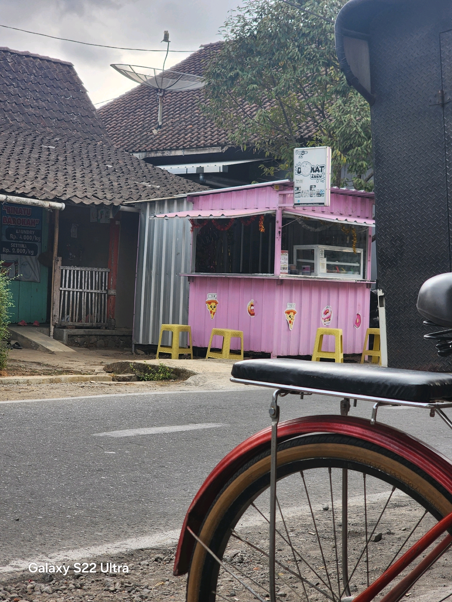 The image captures a scene in Salaman, Indonesia, focusing on a bright pink roadside food stall. The stall, made of a corrugated metal container, is decorated with images of pizza and donuts. Several yellow plastic stools are placed in front of it. To the left, a building with a tiled roof houses a business that appears to be a laundry service, with a sign indicating prices for laundry and ironing. Behind the food stall and laundry, traditional Indonesian houses with tiled roofs are visible. A satellite dish is mounted on one of the roofs. In the foreground, the back wheel of a bicycle is prominently featured, with part of its fender and seat visible. The ground in front of the stall is a paved road, and the overall atmosphere suggests a typical day in a residential or commercial area of the town. The sky is overcast, indicating a cloudy day.