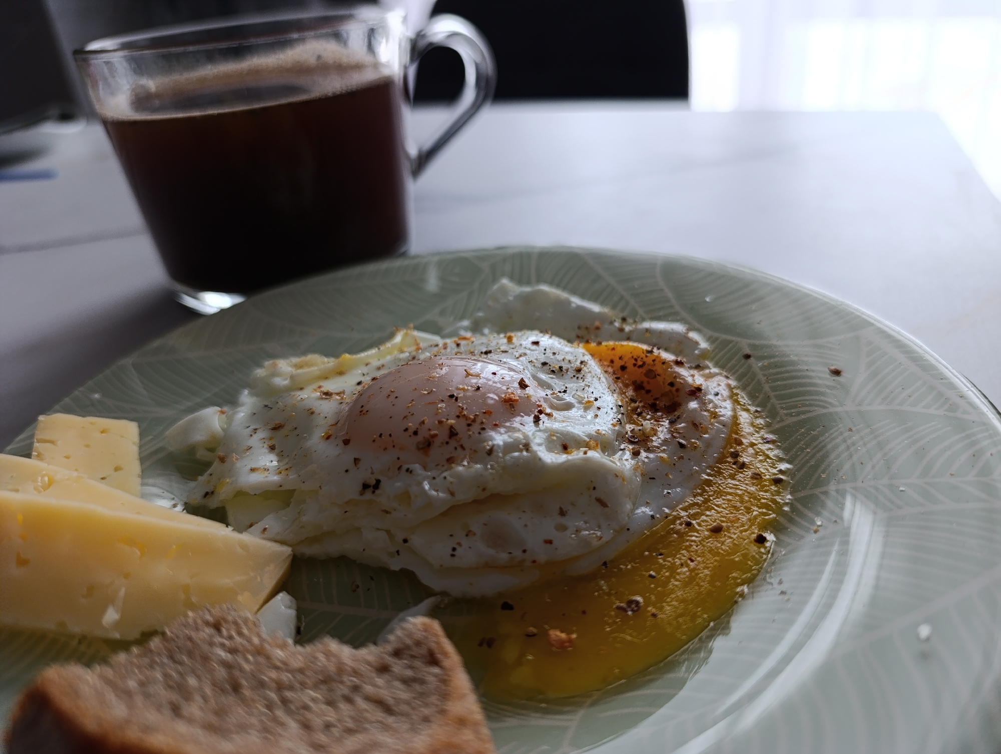 A breakfast scene is captured on a table. In the foreground, a plate is centered, containing a fried egg sprinkled with seasoning, accompanied by slices of yellow cheese and a piece of brown bread. Behind the plate stands a transparent glass mug filled with a dark liquid, possibly coffee. The table surface has a light marble-like pattern. The background includes a blurred glimpse of a window with sheer white curtains. There is no information to indicate this image was taken in the city of Chaikovsky, Russia.