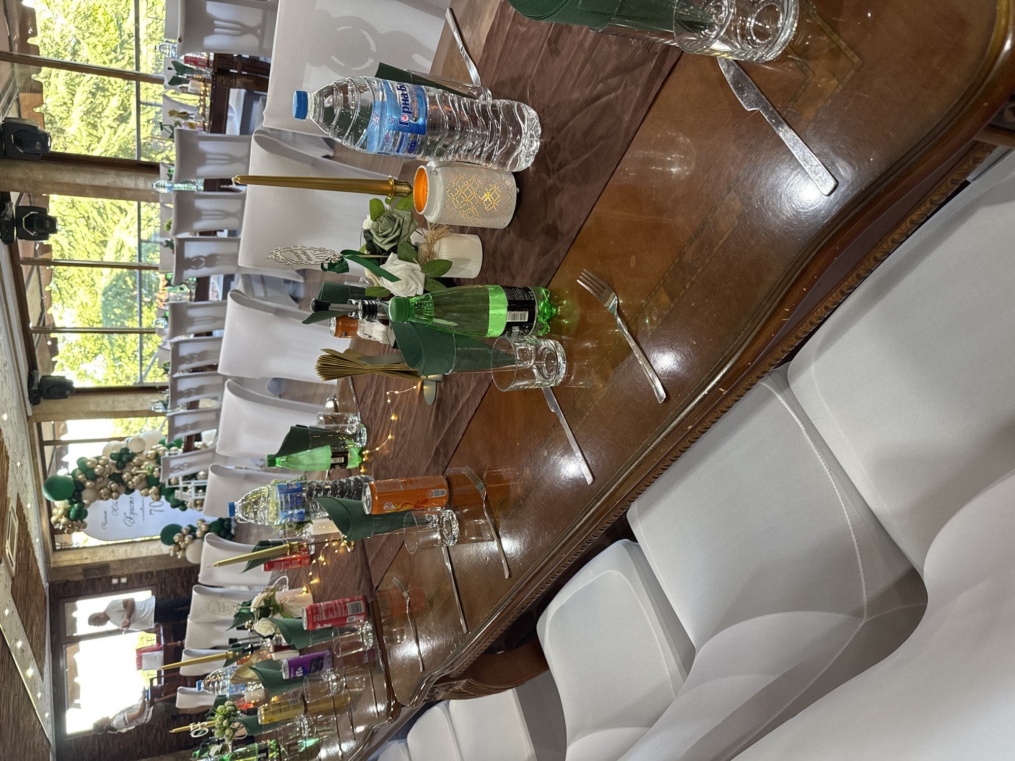 The image depicts a long, polished wooden banquet table set for an event, likely a celebration, in a restaurant or event hall. The table is covered with a dark brown runner, and place settings include silverware, glasses, and green napkins. Several bottles of water and soda are visible, as are decorative elements like candles and floral arrangements. In the background, white chairs are arranged around the table, and large windows offer a view of lush green trees. Two people, an adult male and an adult female, are standing near the entrance area in the background. There is a balloon arch with green and gold balloons, and a banner that appears to be in Bulgarian, possibly indicating a birthday or anniversary celebration with the number "70" prominent. The overall atmosphere suggests a formal yet festive gathering. The location is the restaurant "Borovets" in Sredets, Bulgaria.