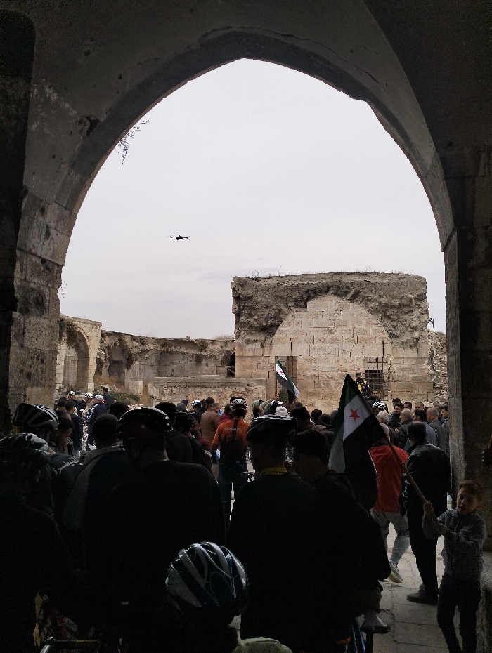 A crowd of people, many wearing bicycle helmets, gathers in what appears to be the entrance of an old, stone structure. Two Syrian flags are visible amidst the crowd. In the sky, a small helicopter is flying in the distance. The stone entrance is arched and leads to a larger open area containing more stone ruins. A young boy is standing towards the right side of the image. The overall scene appears to be in a historic or archaeological site.