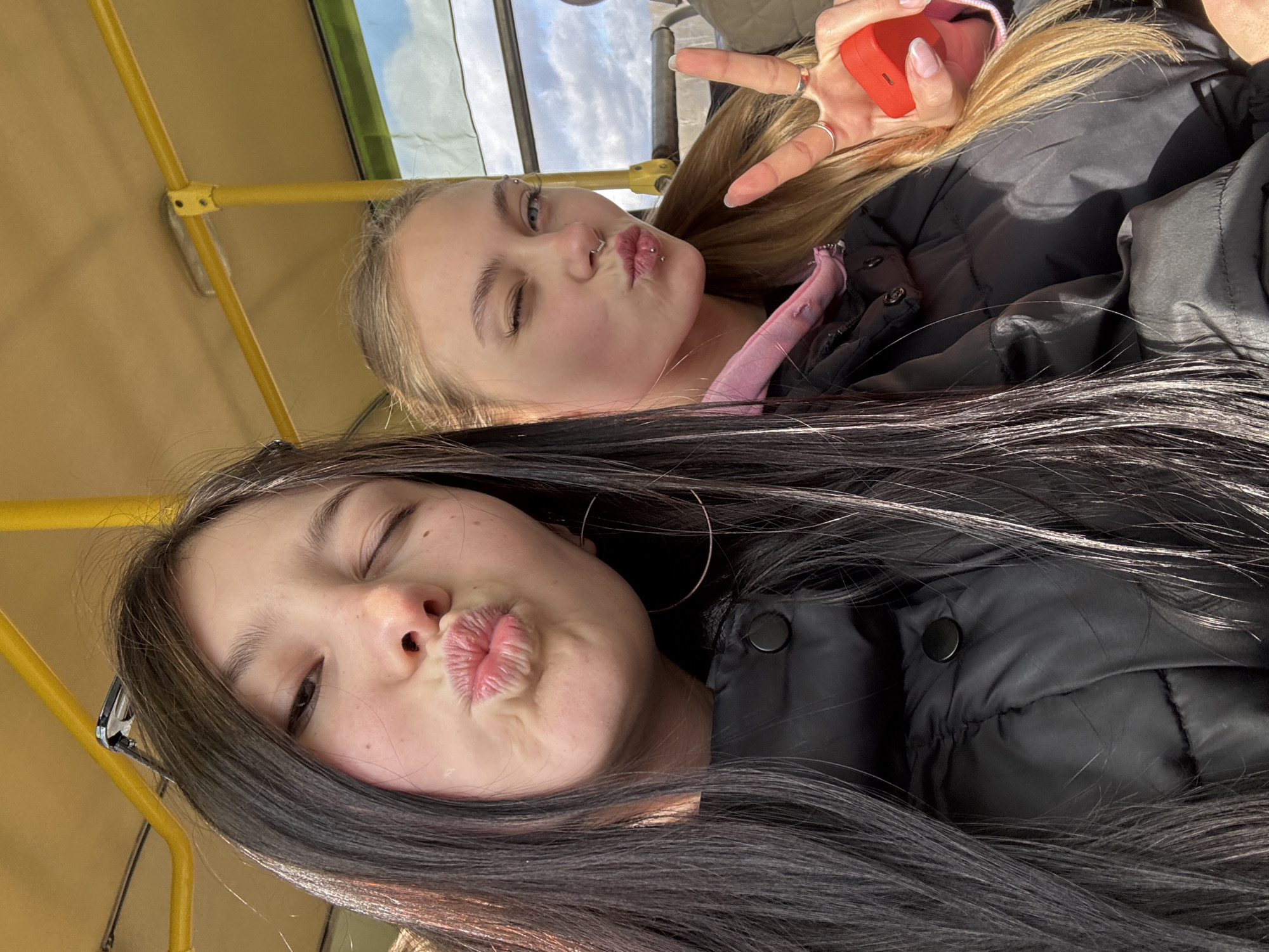This overhead selfie captures two young women inside what appears to be a public transport vehicle, such as a bus or train, identifiable by the yellow metal support bars that frame the top and left sides of the image. Both women are posing playfully for the camera, winking with their right eyes and making "kiss" faces with puckered lips.
The woman in the foreground has long, dark hair flowing over her shoulders and wears a black puffer jacket. A large silver hoop earring is visible on her left ear.
Behind her, the second woman has long, blonde hair and is also wearing a black puffer jacket over a pink garment. She features several facial piercings, including a septum ring and a philtrum piercing. Her left hand is raised, forming a "peace" sign, adorned with rings on her fingers and neatly painted fingernails. She holds a small, red, rectangular object, possibly a phone or a case, in this hand.
Through the vehicle's window, a bright, cloudy sky is visible, indicating it is daytime. The atmosphere of the image is cheerful and friendly, reflecting a moment of shared enjoyment. The scene is situated in Фоки, Russia. There is no visible text in the image.