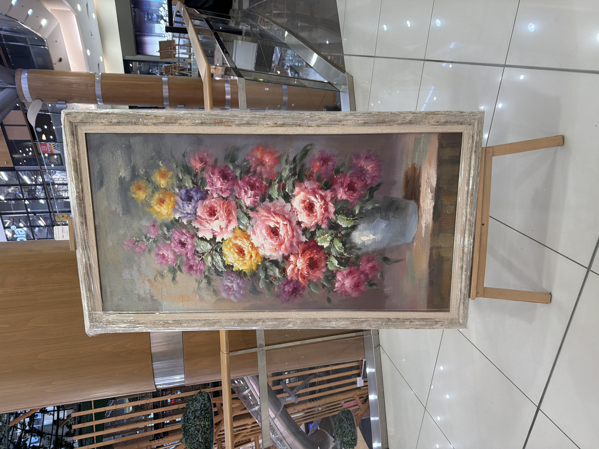 The scene depicts an interior view of a brightly lit commercial building, likely a shopping mall, in the city of Unknown, Unknown. A large, vertical oil painting, depicting a vibrant floral still life, stands centrally on a light wooden easel on a polished white tile floor.
The painting features an abundant bouquet of flowers in shades of pink, fuchsia, yellow, and purple, with green foliage, arranged in a light blue-grey vase. The background of the artwork is a muted blend of grey and brown tones. The painting is encased in a distressed light grey and white wooden frame, which is wrapped in clear plastic film, visible around the edges.
The environment includes large, round pillars clad in light-colored wood with horizontal silver-toned metal bands. A glass and light wood railing system extends diagonally across the midground, suggesting an elevated level or atrium. Reflections in the glass show further architectural elements, including what appears to be a curved slide or escalator and manicured green shrubbery. Distant commercial spaces and additional light wooden easels are visible through the background railings. No individuals are present in the immediate vicinity.