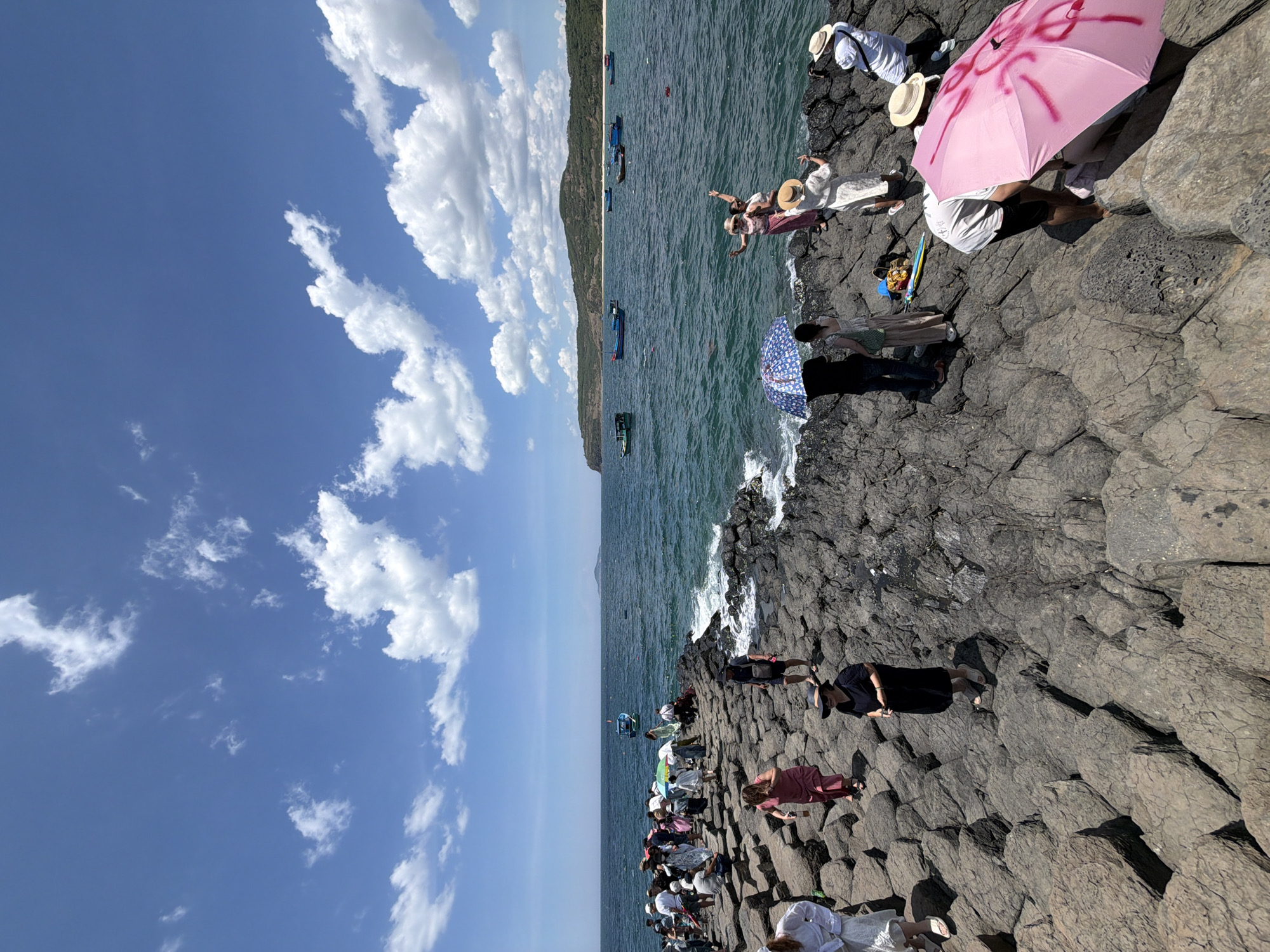 The image depicts a sunny day at a rocky coastal area, possibly in Phú Lương, Vietnam. A group of people are gathered on a formation of dark, hexagonal basalt columns that extend into the sea. The sea is a vibrant blue-green, with waves gently lapping against the rocks. Several small boats are visible on the water in the middle distance, and a hilly coastline can be seen in the background.
The people are dressed in casual summer clothing, and some are holding umbrellas to shield themselves from the sun. A few individuals are posing for photos, while others are walking or standing around, enjoying the view. One person in the foreground is crouching down, and another is holding a pink umbrella with red graffiti on it. The overall atmosphere appears relaxed and tourist-oriented.