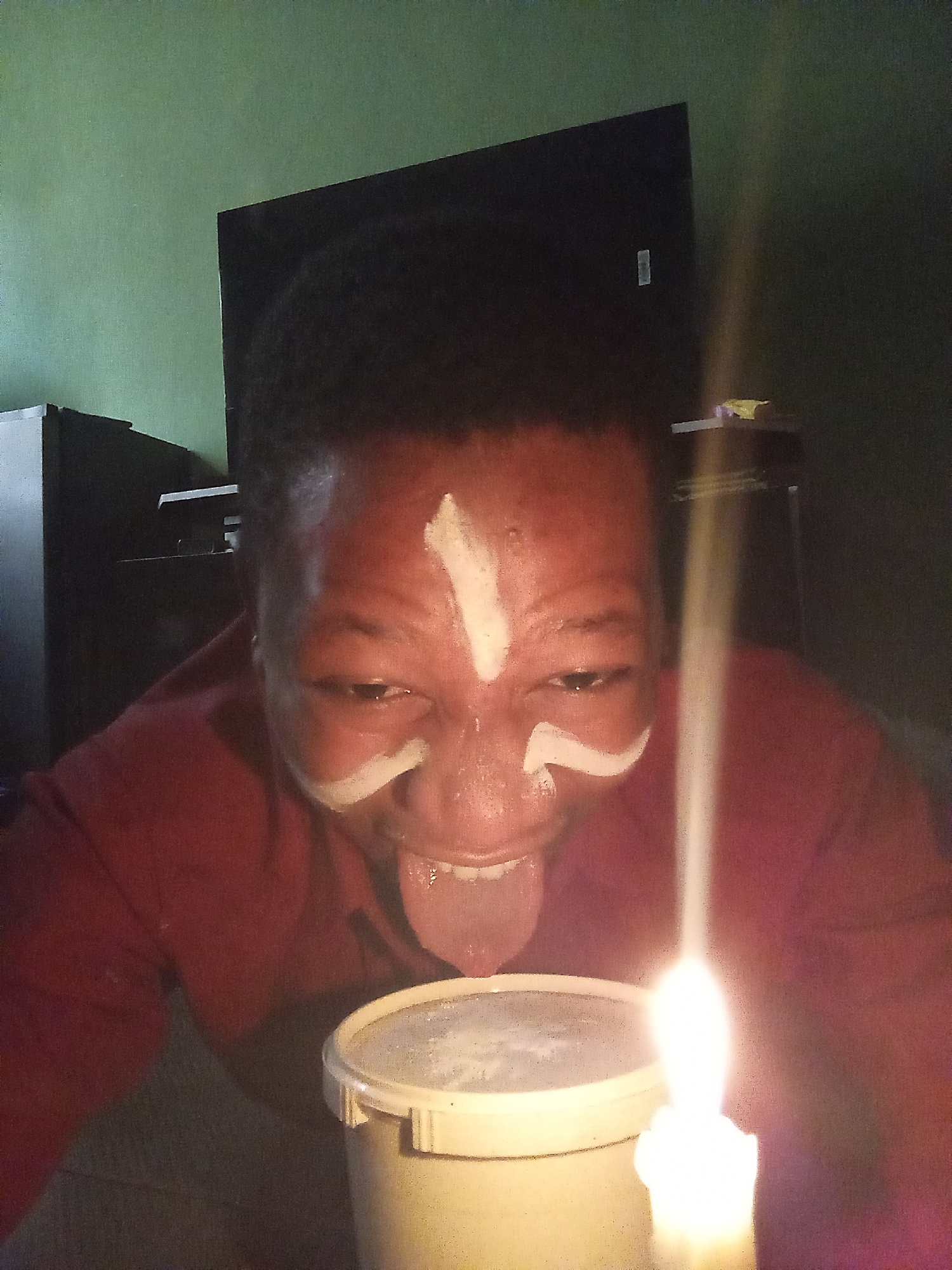A person with dark skin and short, curly hair is positioned in front of a lit candle, their face painted with white markings above the eyes and on the forehead. The person is wearing a red shirt and sticking out their tongue towards a white container with a lid. Behind the person are various dark-colored furniture pieces, including a television, against a dark green wall. Based on the attire and markings, the person's appearance aligns with cultural practices in the Oshogbo region of Nigeria.