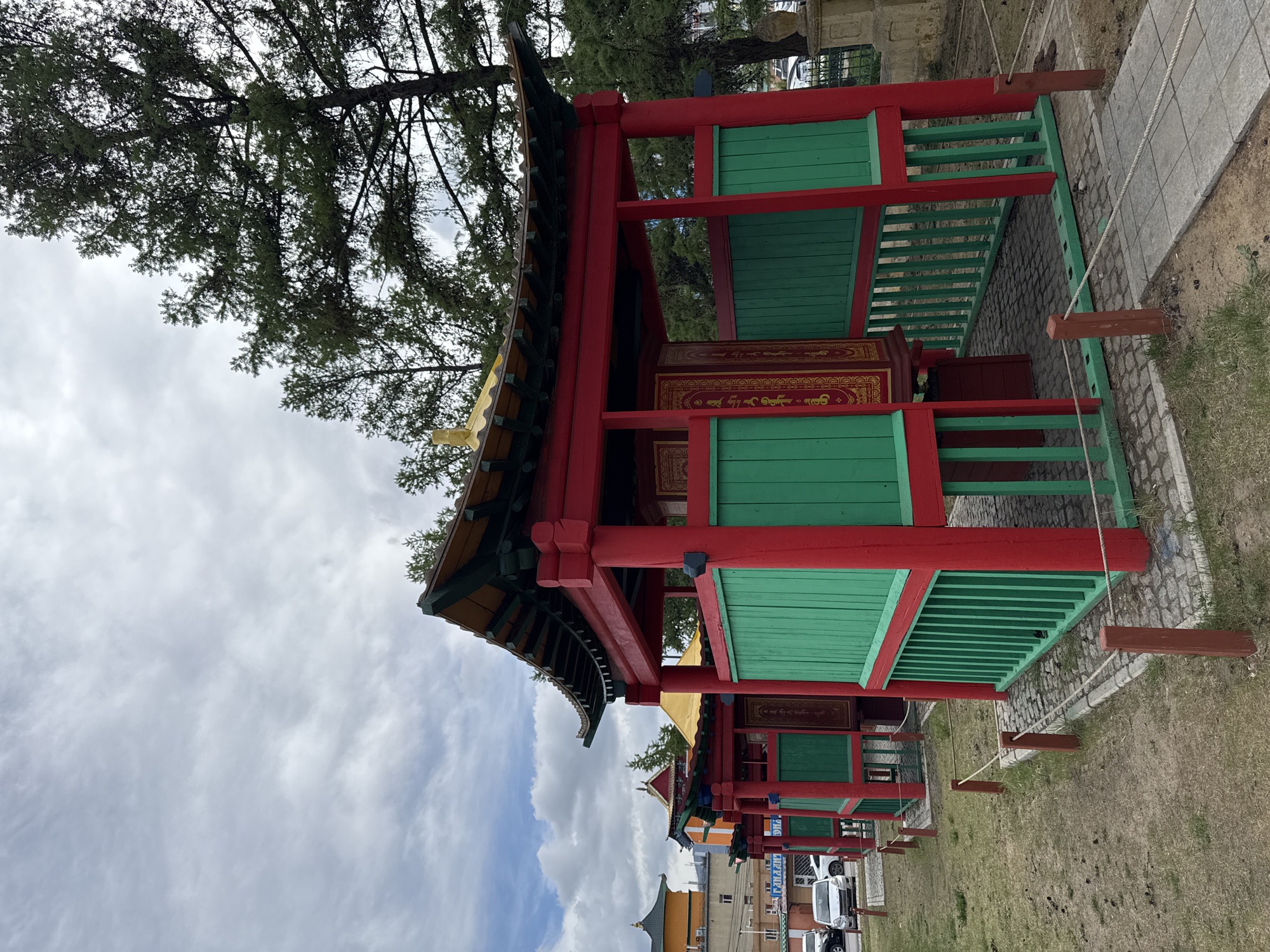 A low-angle shot depicts a series of small, elaborately decorated red and green structures under a cloudy sky in Ulaanbaatar, Mongolia. The structures feature ornate roofs with detailed carvings and green latticework. The red paint is a primary color, contrasting with the light green. Each structure has a painted interior and is linked by a rope fence with evenly spaced wooden posts. The background includes a tall tree and modern buildings, contrasting the traditional structures. Pedestrians are visible in the background, and the surrounding ground is a mix of paving stones, cobblestone, and grass.