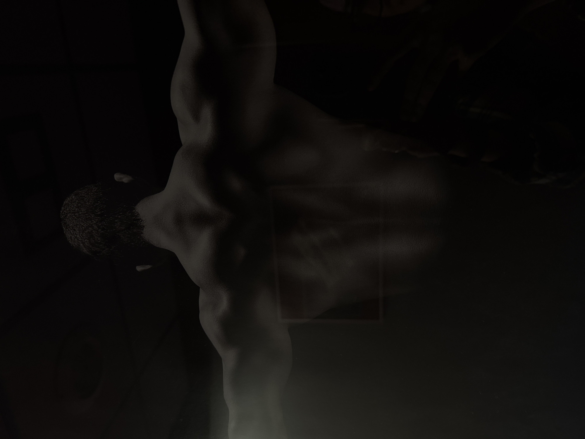 The image shows the back of a shirtless man with muscular definition, facing away from the camera. His arms are extended outwards. The lighting is dramatic, with shadows obscuring much of the background and parts of the man's body. A faint, rectangular outline is visible on his back. The overall setting appears to be indoors, possibly a gym or a dimly lit room, with a dark ceiling. There is no identifiable location in Riga, Latvia, present in the image.