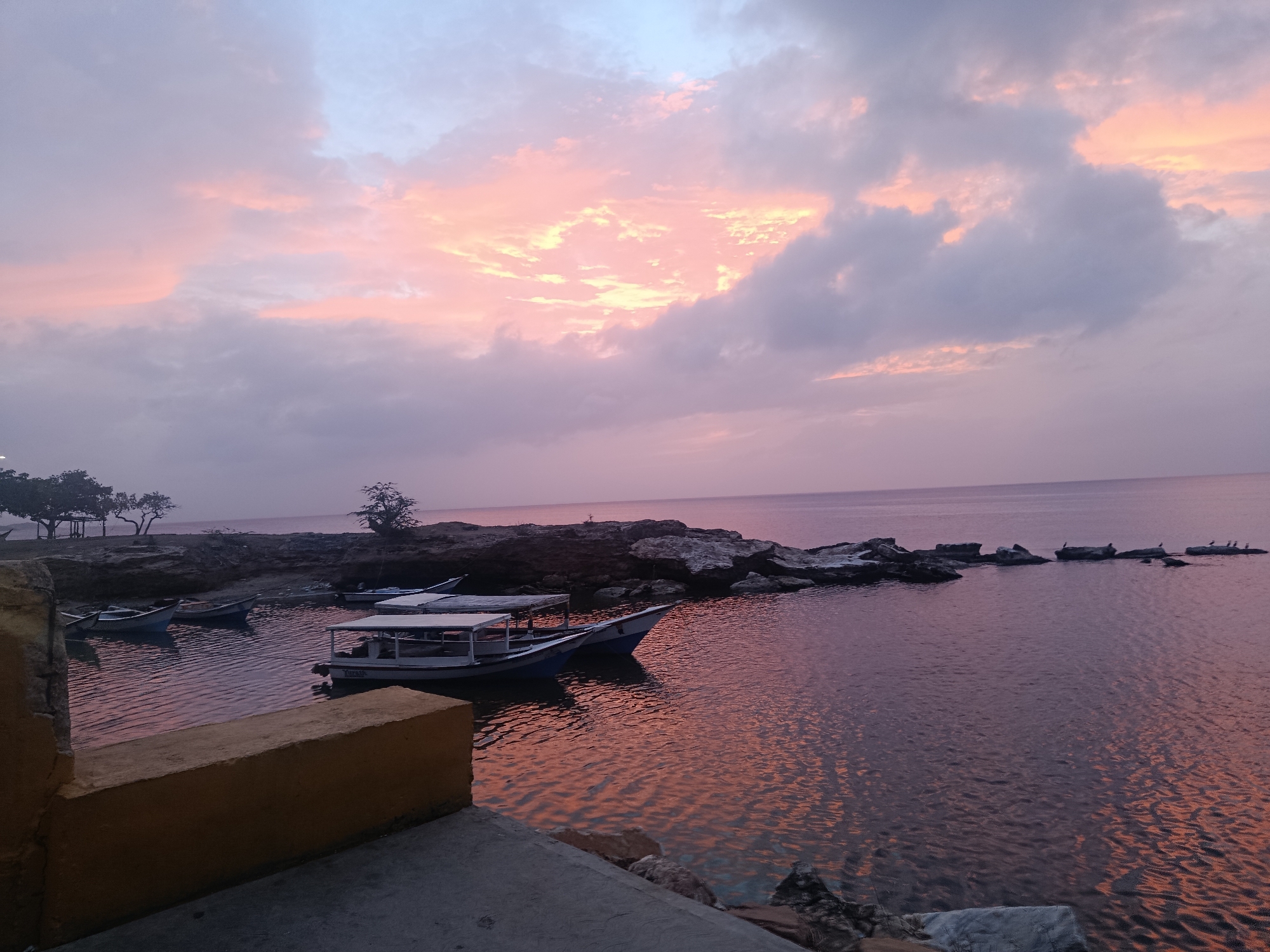 The image shows a bay scene at sunset in Juan Griego, Venezuela. Several small boats are docked near a rocky shoreline. The sky is filled with pink and gray clouds, reflecting in the water. Rocky outcroppings extend into the sea in the distance. A low concrete wall is in the foreground.