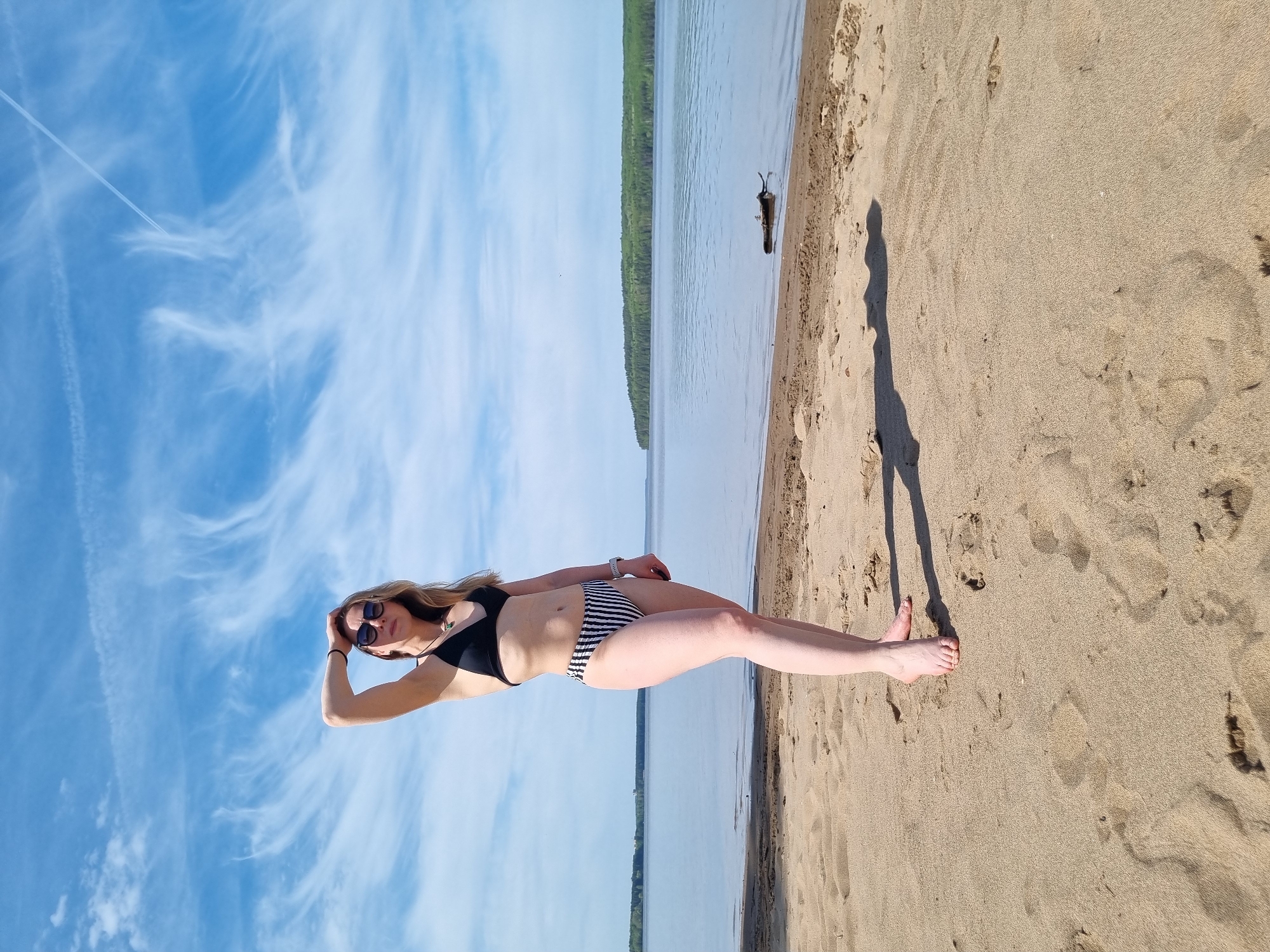 A woman is standing on a sandy beach near a body of water, possibly in the city of Chaikovsky, Russia. She is wearing a black bikini top and striped bikini bottoms. She is wearing sunglasses and has her hand on her head. The sky is blue with wispy clouds, and there are trees visible across the water. A piece of driftwood is floating in the water near the shoreline. The woman's shadow is cast on the sand.