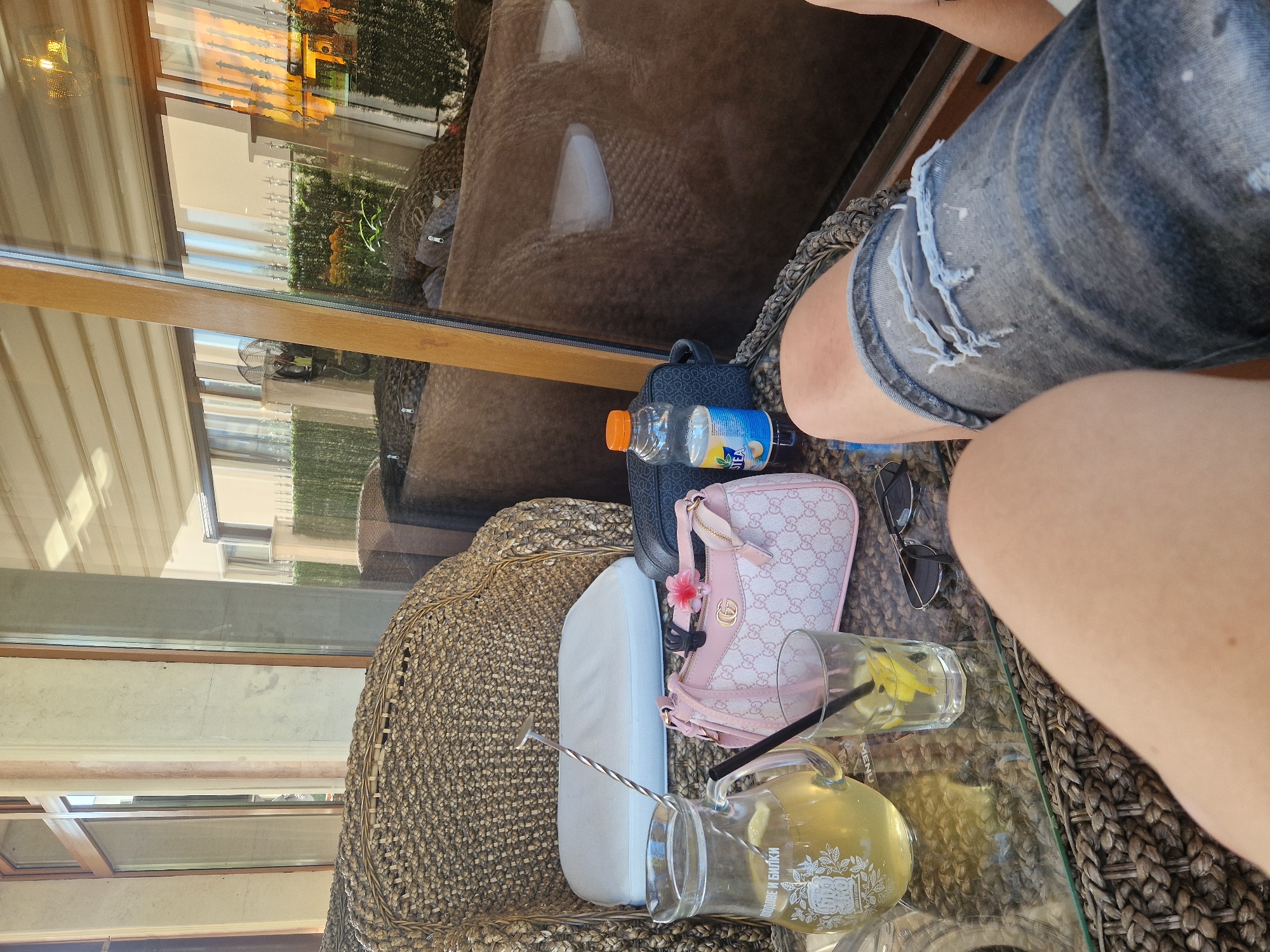 The image depicts a close-up view of a table in an outdoor seating area, likely a cafe or restaurant, situated in Sofia, Bulgaria. A person's legs, clad in ripped denim shorts, are prominently featured in the foreground. On the table, a glass pitcher filled with a light-colored beverage and lemon slices sits next to a glass with a straw and lemon. A pink designer handbag with a prominent "GG" logo and a floral embellishment is placed to the right of the pitcher. Also on the table are a bottle of iced tea, a pair of sunglasses, and a bar spoon. The background shows a wicker chair and a reflective glass surface that offers a blurred view of the surroundings, including outdoor seating and greenery, suggesting an urban or resort-like setting. The sunlight and shadows indicate it is daytime.