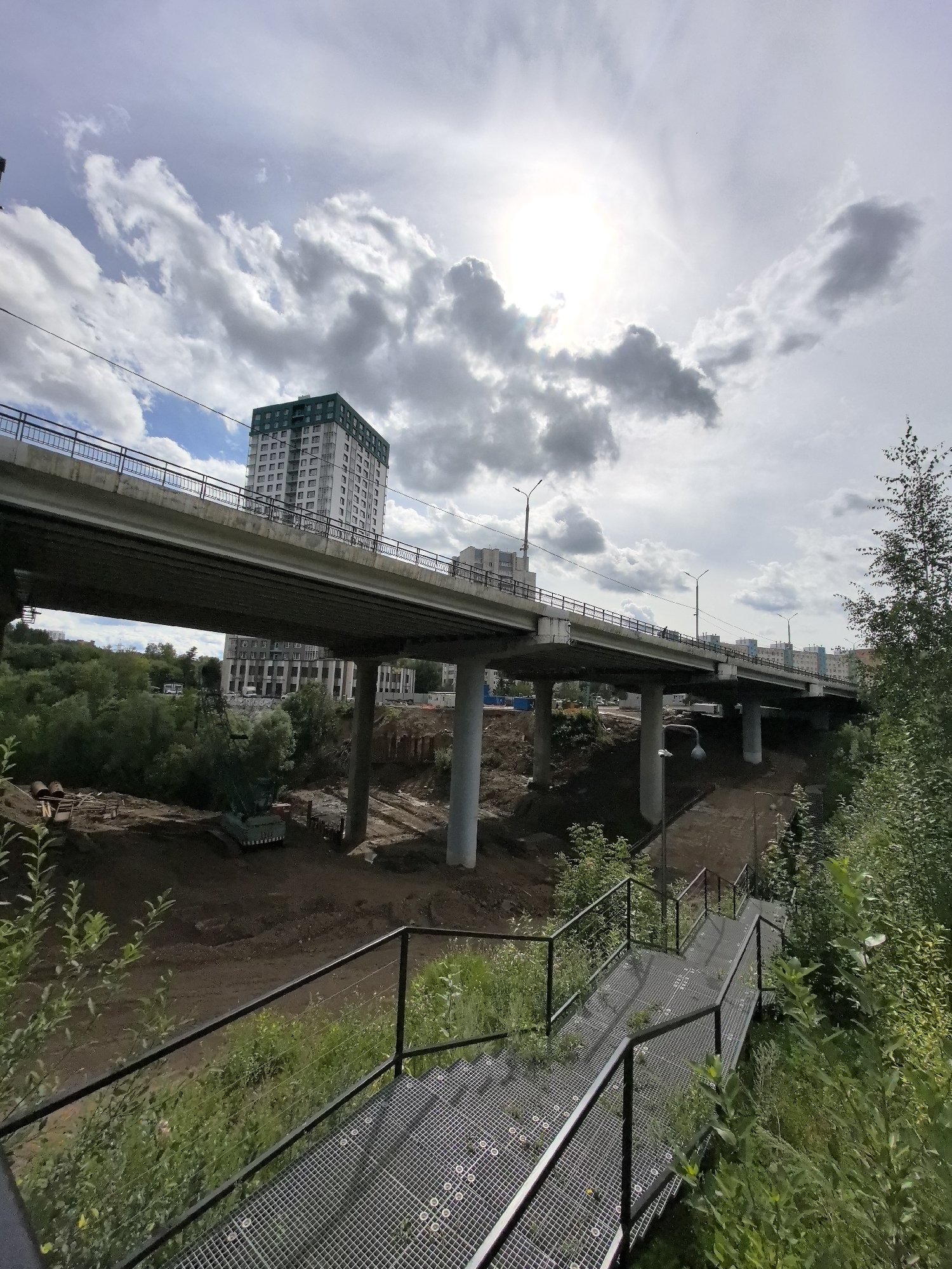 The image depicts an elevated highway or overpass under construction in Perm, Russia. The overpass spans across a low-lying area with a construction site visible below, featuring a crane and disturbed earth. A multi-story building with a distinctive green roof is situated in the background, to the left of the overpass. Another building, also appearing to be residential, is visible further in the distance. The foreground shows a metal staircase leading downwards, with lush green vegetation growing around it. The sky is partly cloudy with the sun peeking through, casting a bright glare in the upper center of the image. The overall scene suggests ongoing urban development or infrastructure improvement.