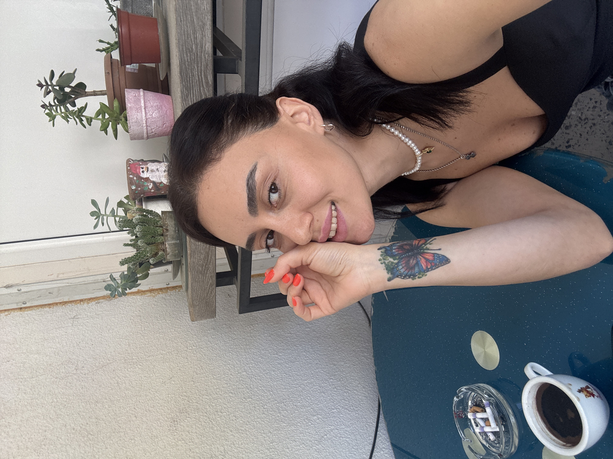 The image shows a woman with dark hair and fair skin, wearing a black tank top and a pearl necklace. She has a butterfly tattoo on her left forearm and bright orange nail polish. She is smiling and looking towards the camera. In the foreground, there is a dark blue table with a white coffee cup and an ashtray containing several cigarette butts. Behind the table, there are several potted plants on a shelf, including succulents and a cactus. The background appears to be an outdoor setting, possibly a balcony or patio, with a textured wall and a white door or window frame. The overall scene suggests a casual moment outdoors, perhaps in Tbilisi, Georgia, given the provided context.