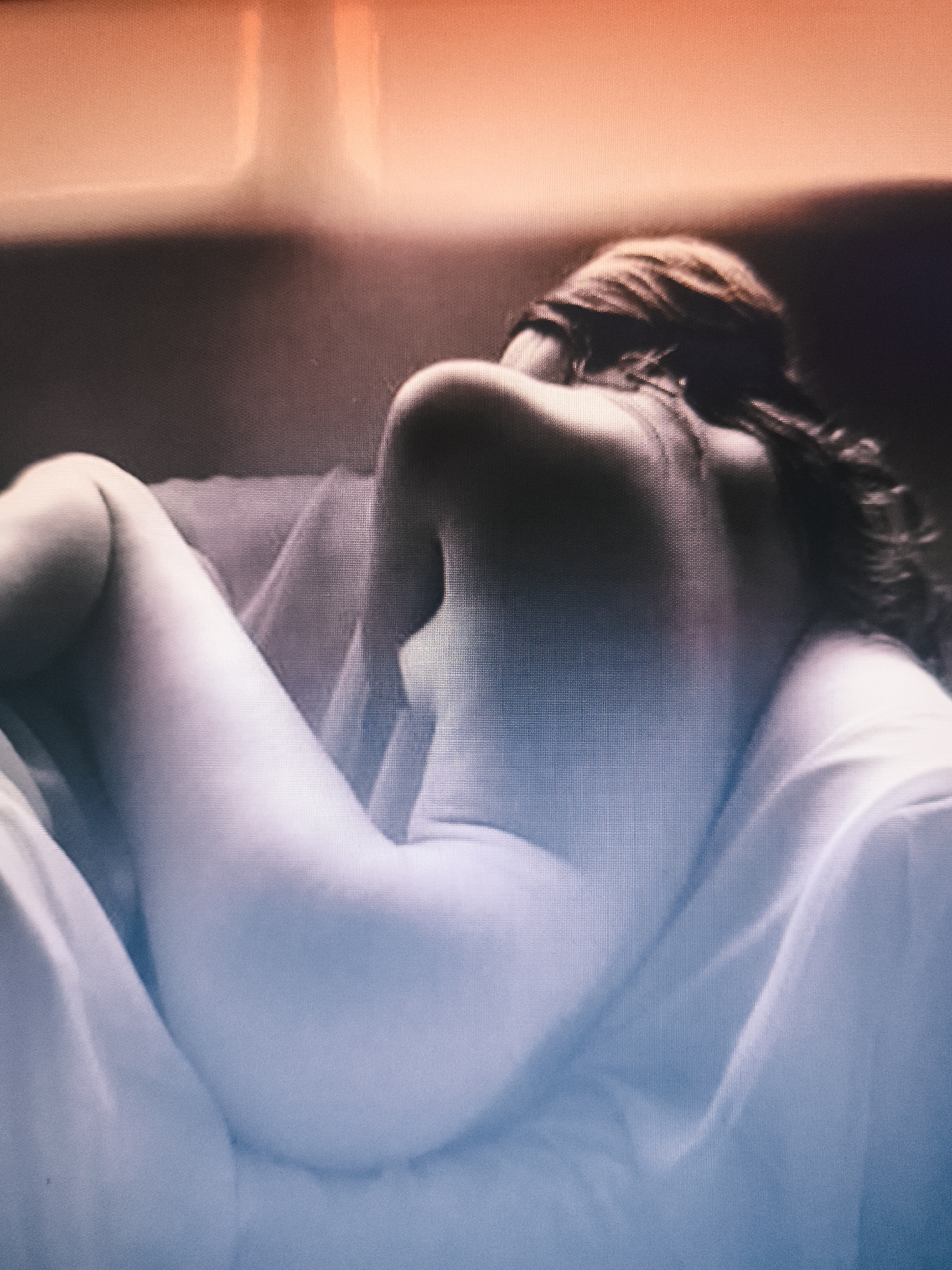 A woman is reclining on a white draped surface. The image is cropped so that only the woman's back, torso, and legs are visible. She appears to be facing away from the viewer, with her head resting on something. Her hair is long and flowing. The image has a gradient effect, with the top half being a warm, orange tone and the bottom half a cool, blue tone. There is no indication of this image being in Nowy Tomyśl, Poland.