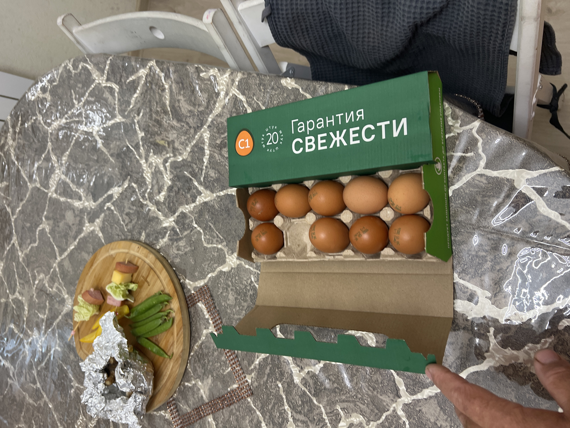 An overhead medium shot captures a tabletop scene featuring a carton of brown eggs and a wooden platter of appetizers. The tabletop appears to be covered with a plastic tablecloth that mimics a marble pattern.
In the foreground, to the left, a person's hand is partially visible, with fingers extended towards the edge of the frame, suggesting they are interacting with the scene.
To the right, a green cardboard carton is open, revealing several brown eggs nestled within its molded compartments. The carton has text printed on it in white lettering. The visible text includes "Гарантия свежести" (guarantee of freshness), and other markings that suggest quantity and quality. Some of the eggs have stamps on them, which are difficult to decipher in detail but appear to contain dates.
To the left of the egg carton, a round wooden platter holds various food items. On the platter, there are some green beans, a piece of food wrapped in aluminum foil, and several small skewers. Each skewer contains a combination of what appear to be chunks of cheese, sausage, and possibly lettuce or other greenery. The arrangement suggests a casual appetizer setting.
The background is slightly out of focus, but a white chair is partially visible on the right side of the image. The overall impression is of a domestic or casual dining setting. The specific mention of Sochi, Russia, in the prompt does not have any visual cues within the image to confirm or deny this location.