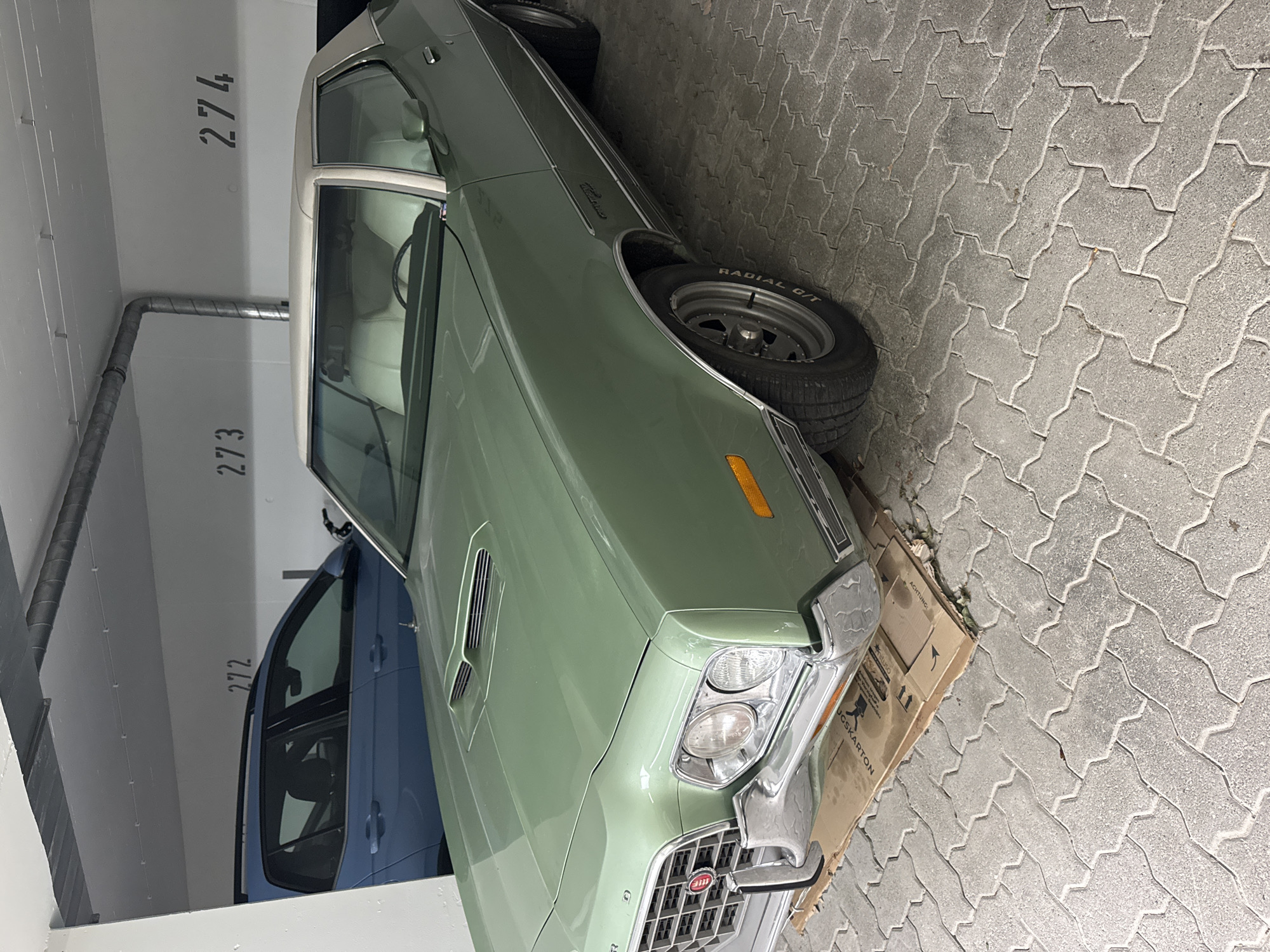 The image captures a green classic car, likely a 1970s Ford Gran Torino, parked in an underground parking garage. The car is the primary subject, with its light-colored roof and interior visible. It is positioned on a section of paver bricks, with a cardboard mat placed underneath the front of the vehicle. In the background, a blue car is parked to the left, and parking spot numbers "272", "273", and "274" are stenciled on the white wall. The environment appears to be an indoor parking facility with overhead pipes and concrete walls. The image's metadata indicates it was taken in Höhenkirchen-Siegertsbrunn, Germany, on August 23, 2022. No people are present in the image.