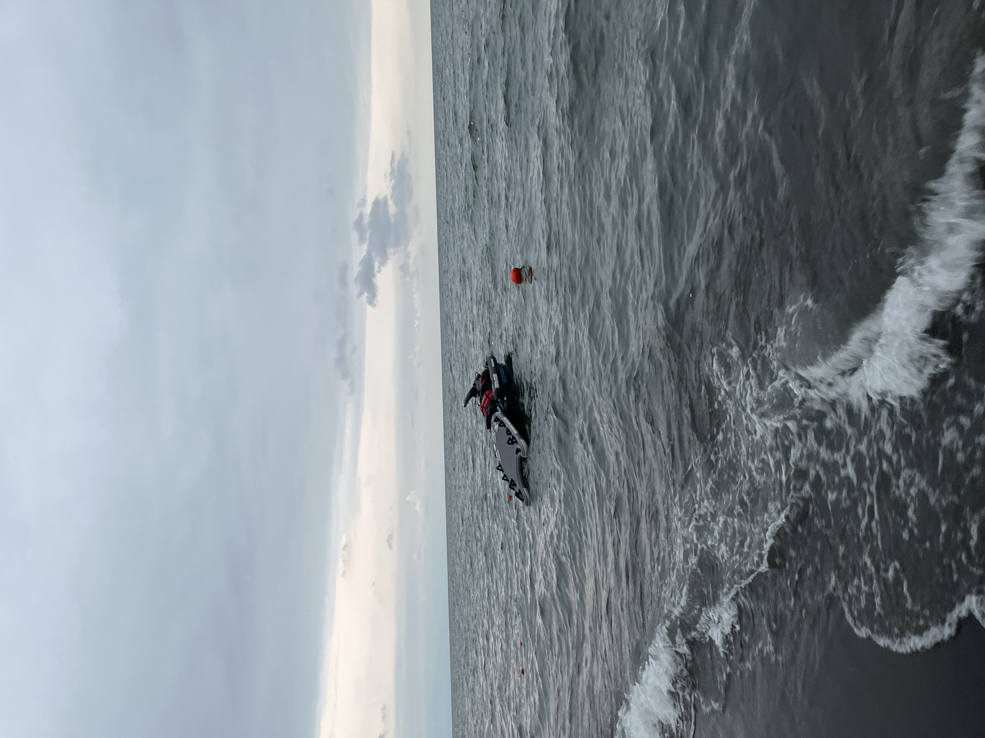 A jet ski is partially submerged in the choppy, dark grey waters of the Black Sea near Ureki, Georgia. The jet ski, a black Sea-Doo model, is tipped to the side, with its front partially underwater. A dark grey platform, possibly a jet ski dock or a wakeboard, is attached to the rear of the jet ski and is floating on the surface. A bright orange buoy is visible in the water to the right of the jet ski. In the distance, a net or string of buoys stretches across the water. The sky is overcast with grey clouds, and the sea is a deep grey, with small waves breaking on the dark sand shore in the foreground. No people are visible in the image.